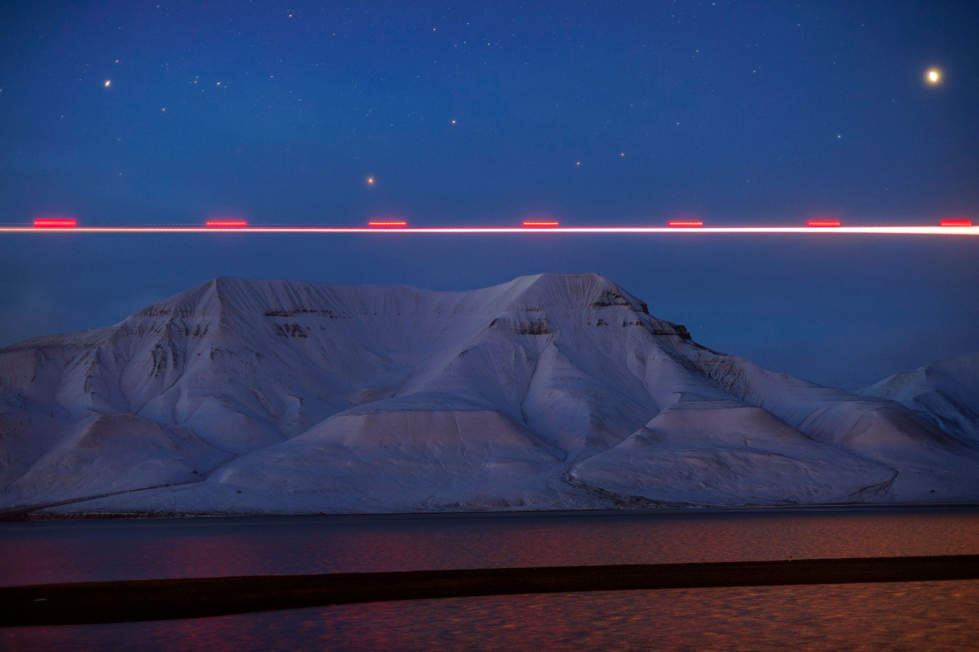 Horizontal red light trail, caused by a plane, reflecting in the fjord as it passes a mountain