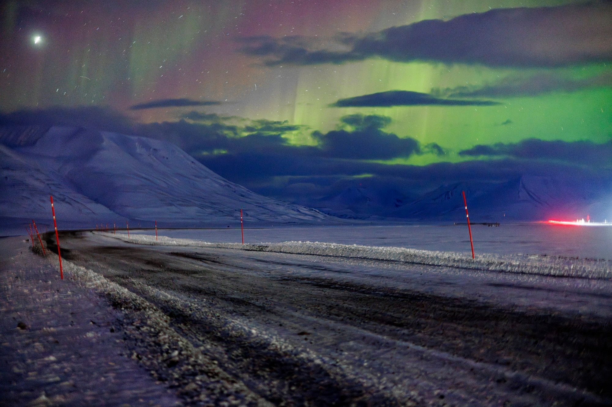 The aurora borealis visible over a snow covered mountain with a road in the foreground and a small glow from a car's brake lights in the background