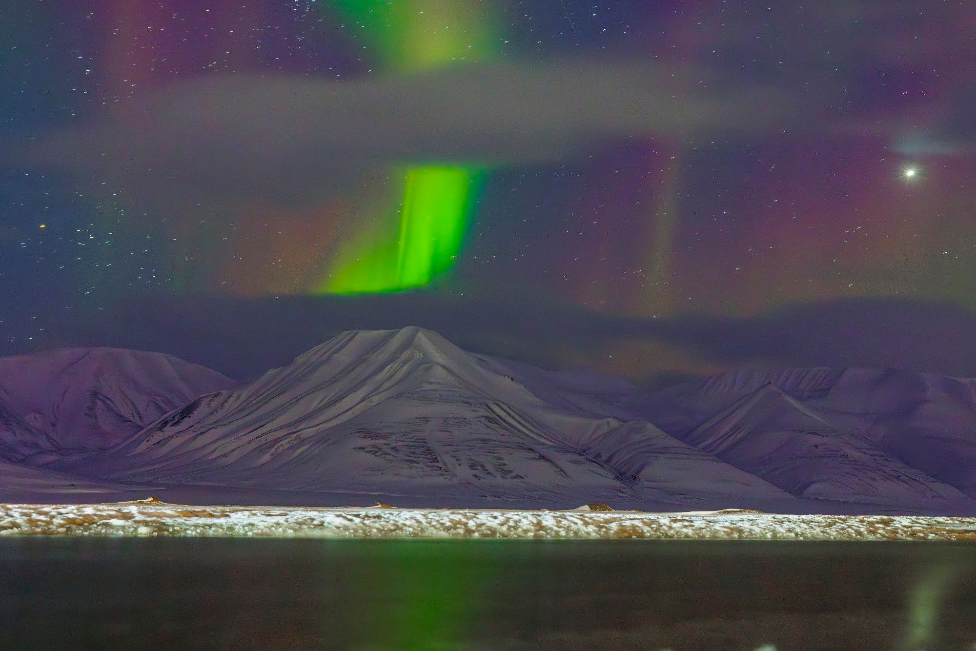 The aurora borealis visible over a snow covered mountain with a faint reflection on some water in the foreground