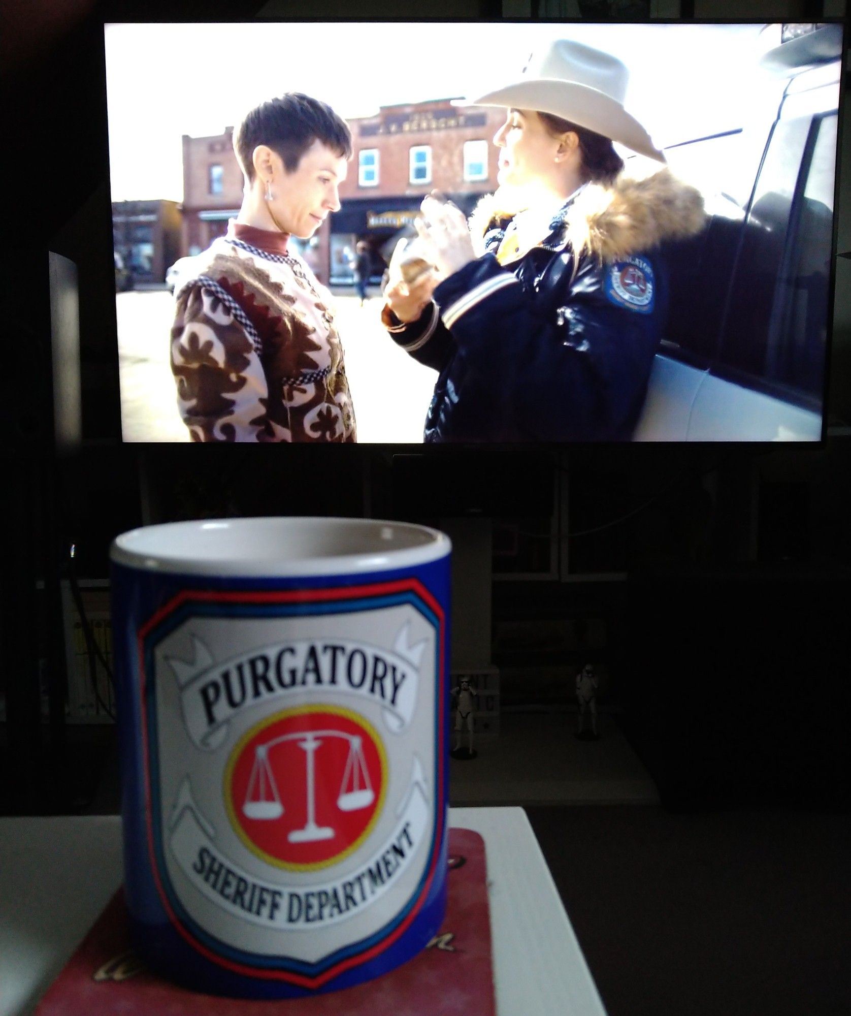 Scene from Wynonna Earp Vengeance playing on a TV with a Purgatory Sheriff Department mug in the foreground.