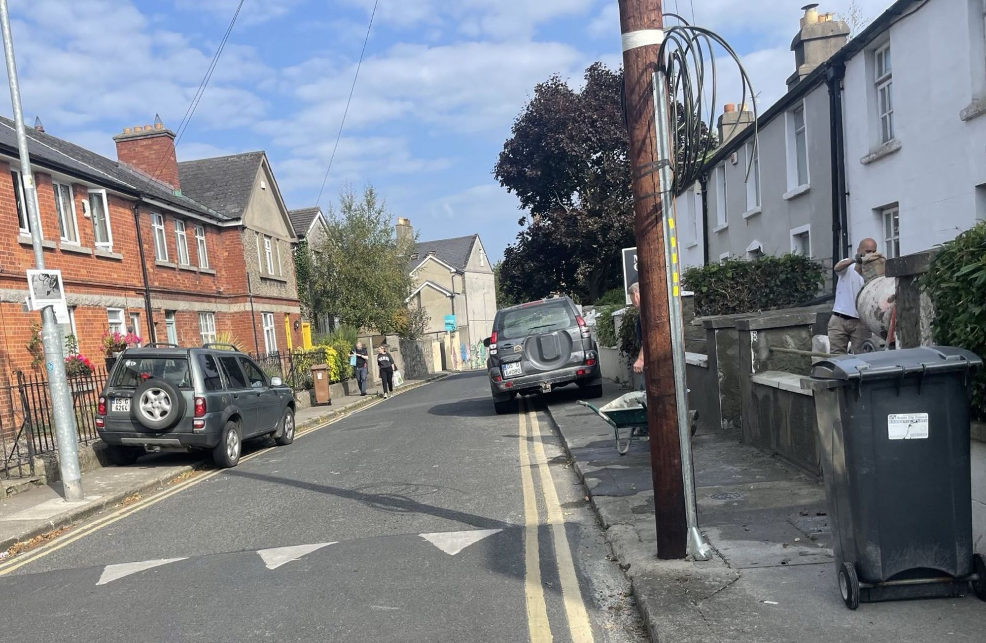 Mount Pleasant Avenue, Dublin 6, with two grey jeeps, each one parked mostly on the footpath, one on each side of the road. There's a wheelbarrow on the footpath on the right side, and a builder operating a small concrete mixer in the garden of a terrace house.