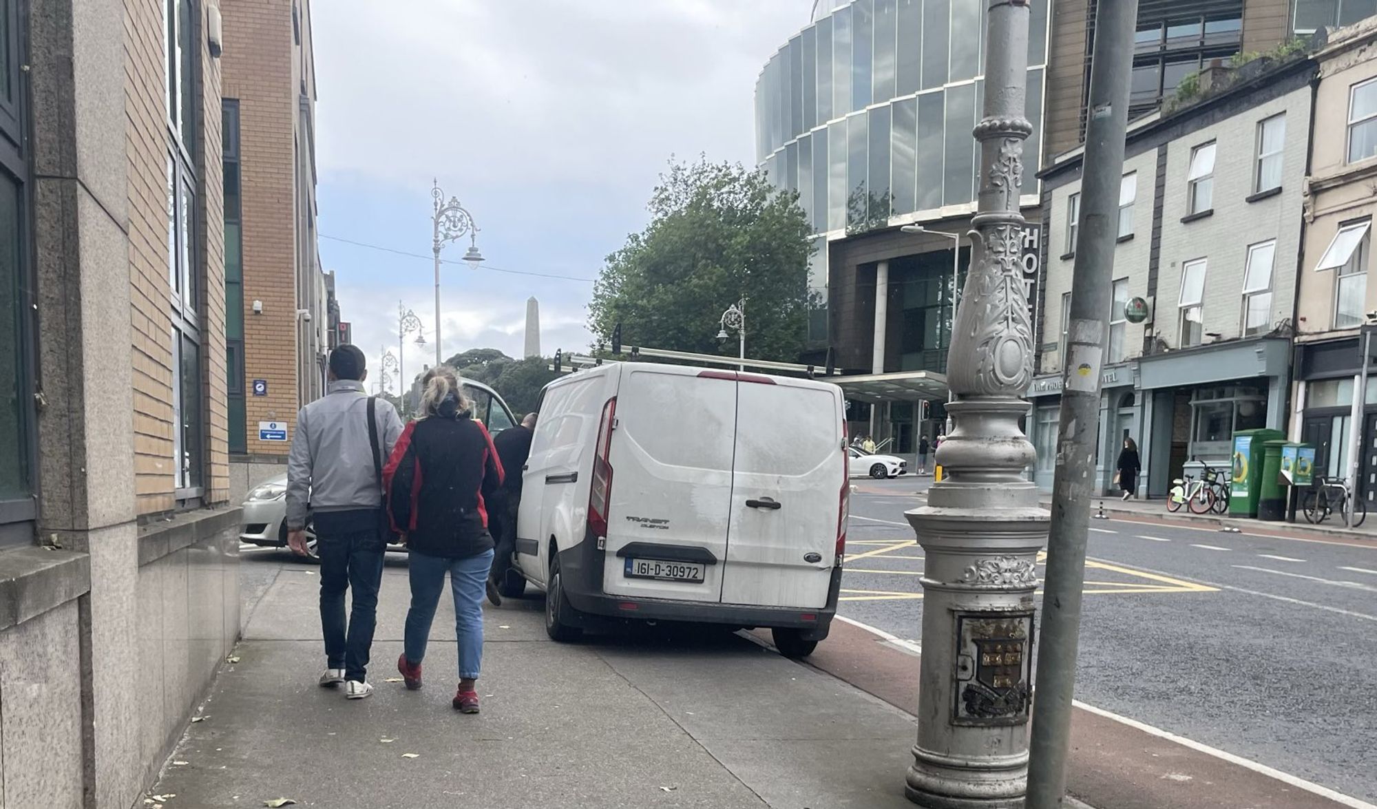 Parkgate St, Dublin 8 - white Transit style van parked mostly on the footpath, with the outer wheels sitting in the cycle lane. The passenger door is open and someone is climbing into the van. There's a couple walking on the remaining part of the footpath.