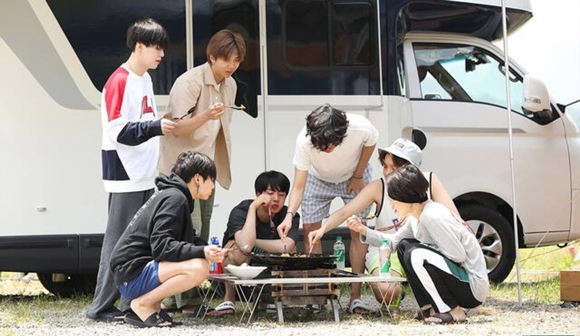 Members eating outside, in front of a van.
Some of them are standing, the others are crouching.
They are gathered around a caldron and eat together.