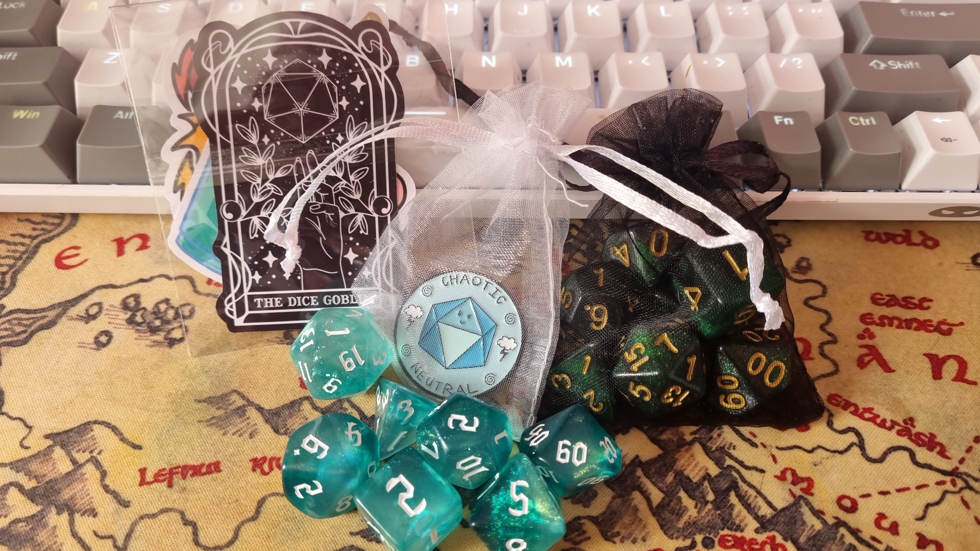 a picture of two sets of dice, a light blue glittery set, a dark green glittery set, a pack of assorted D&D related stickers and  a pin that's says "chaotic neutral" with a d20 on it