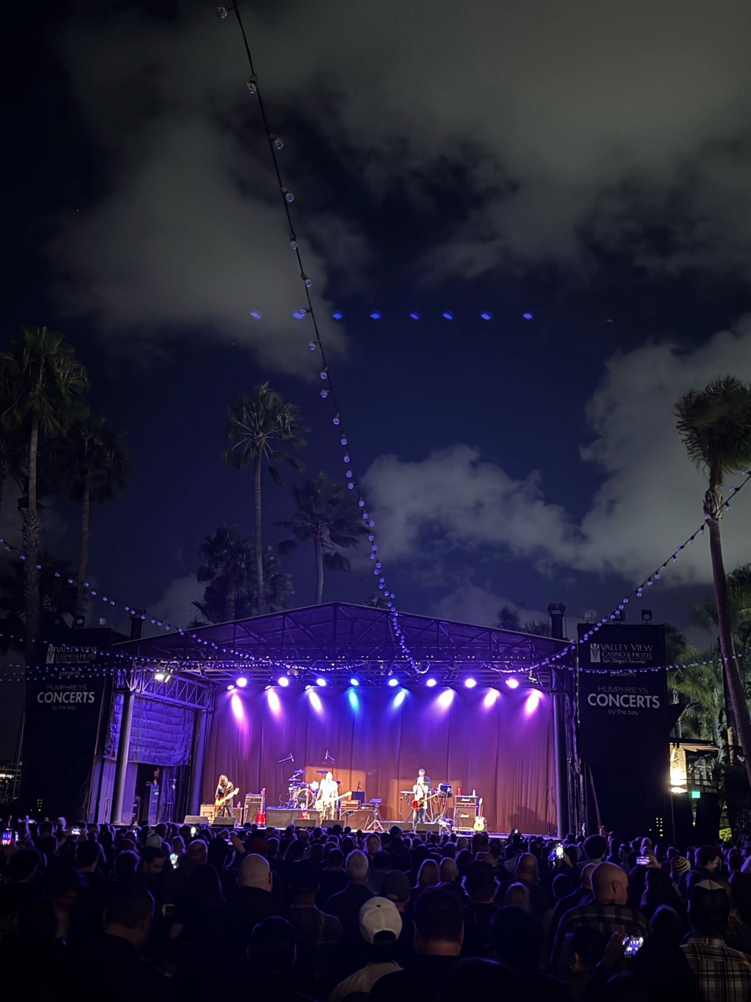 Wide shot of Peter Hook and band on stage at Humphreys in San Diego. Palm trees and clouds behind.