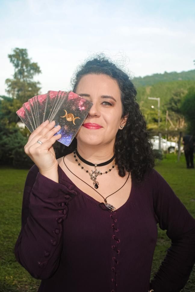 Mulher branca de cabelo escuro cacheado com vestido medieval roxo, segurando um deck de tarô na frente do rosto. Ao fundo, um gramado verde, árvores, cenário bucólico.