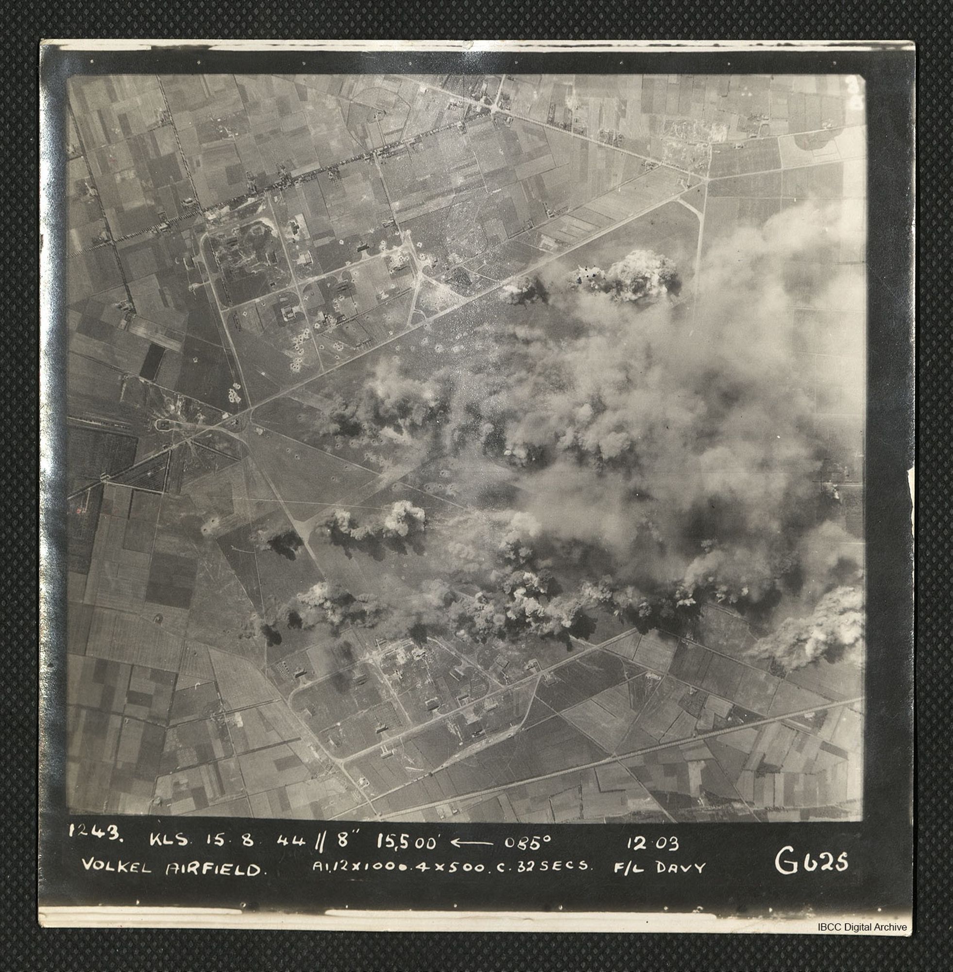 A vertical aerial photograph of an operation at Volkel airfield. Much of the image is obscured by bomb blasts and smoke.
It is captioned '1243 KLS 15.8.44//8" 15,500 <-- 085° 12.03 Volkel Airfield A1 12x1000. 4x500 c. 32 secs. F/L Davy G625'.
IBCC Digital Archive item 43513