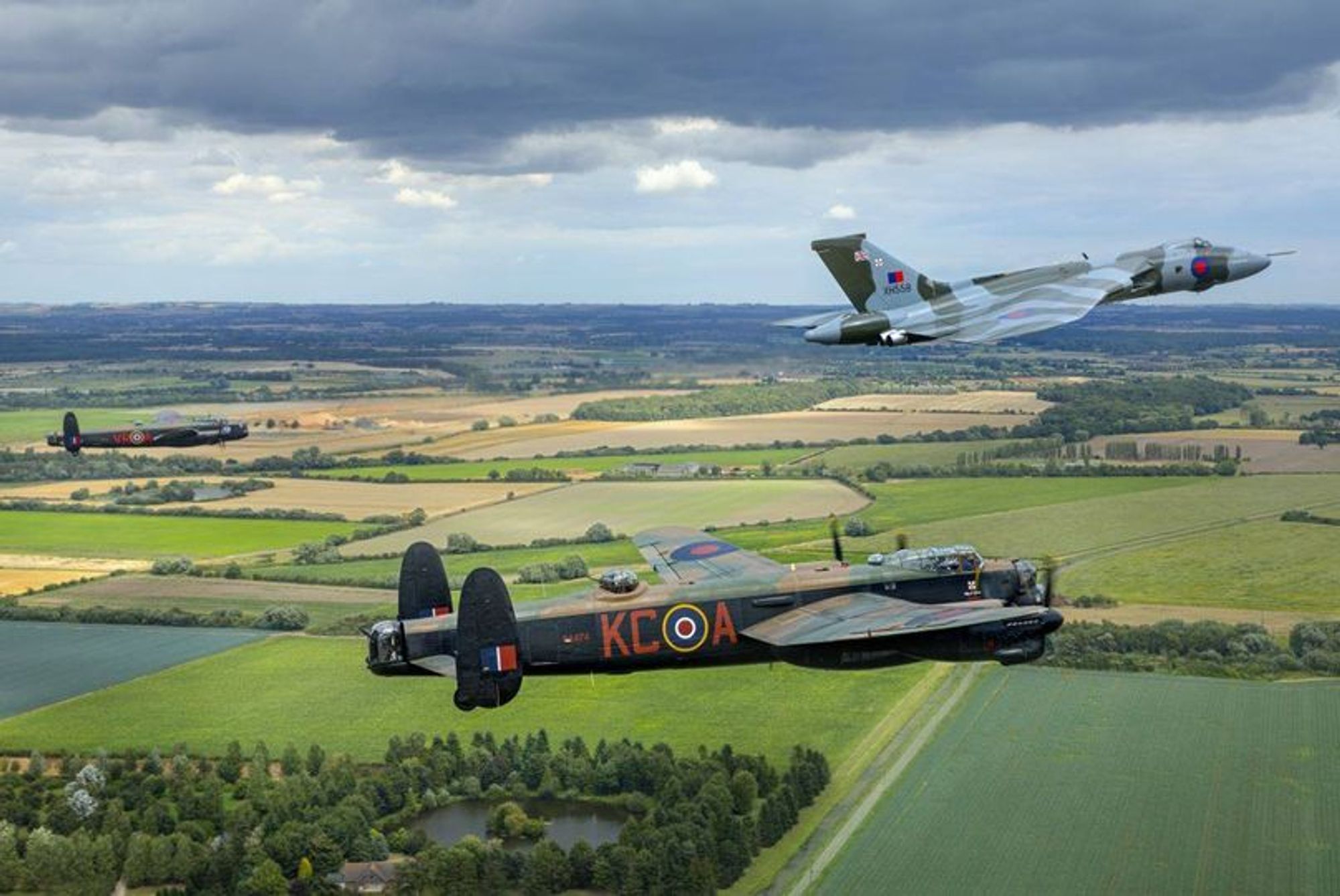 Air-to-air photo of the two Lancasters and the Vulcan in formation over patchwork fields.