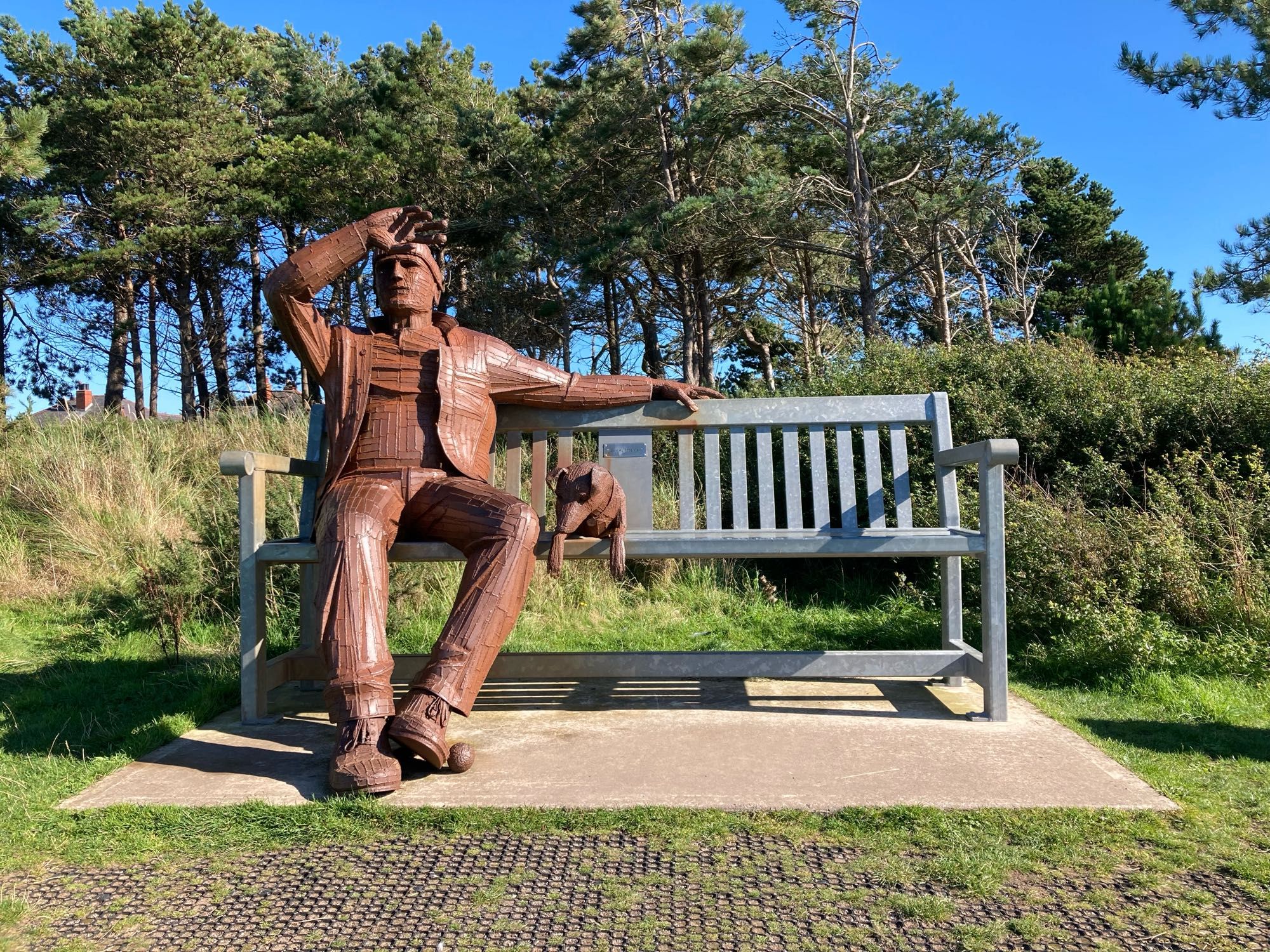 Bigger than lifesize metal sculpture of man on an equally big park bench style seat. He has a ball at his feet and his dog is sitting next to him with eyes focussed on the ball.