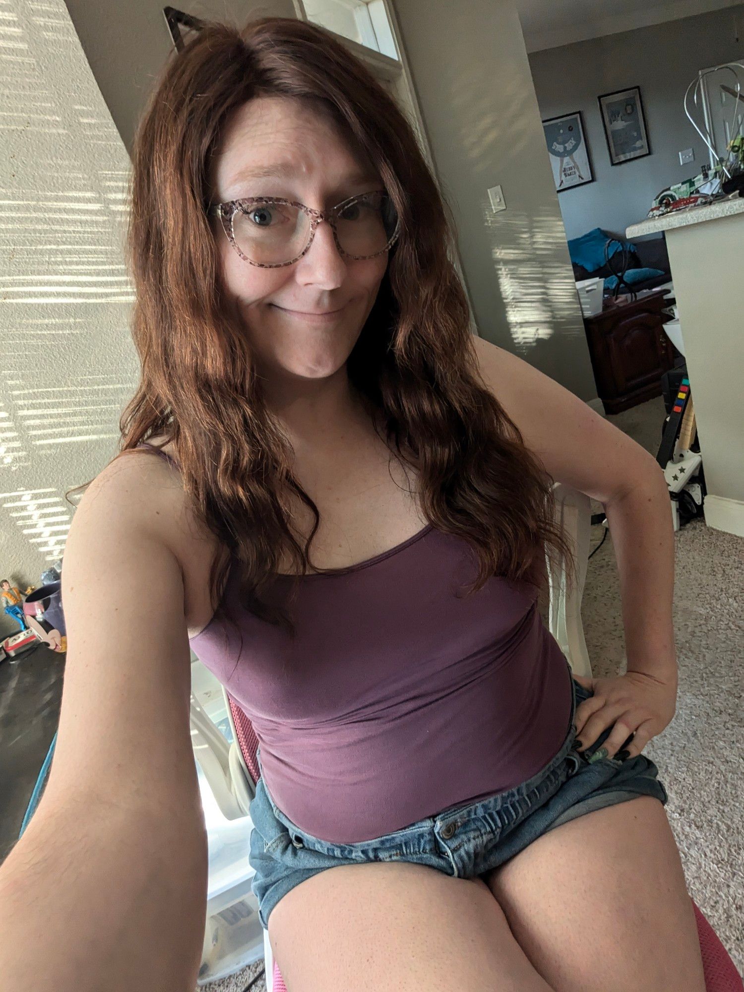 Janey sits on her pink office chair smiling, wearing her trademark brown floral glasses along with a purple camisole and short denim shorts.