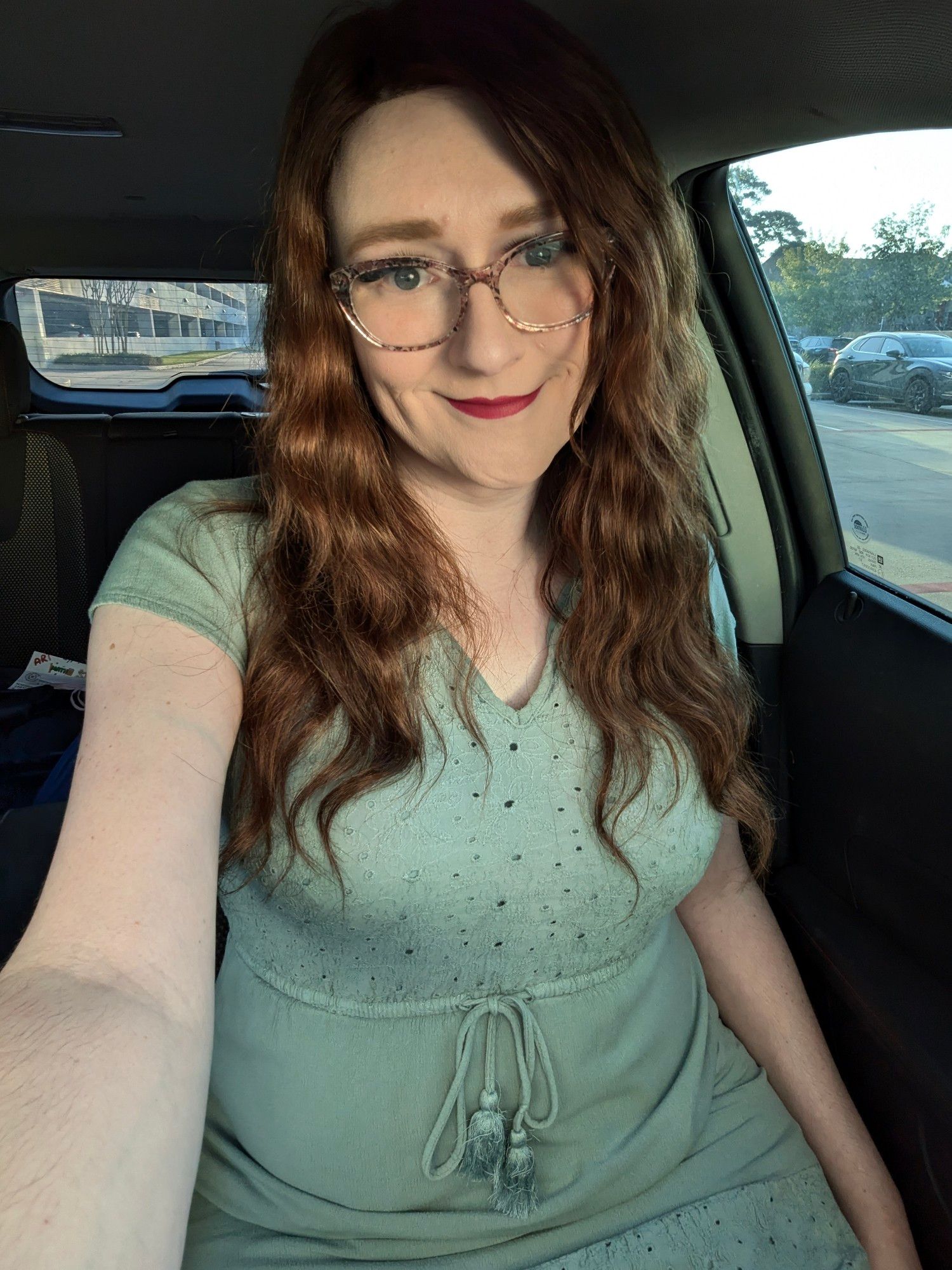 Janey sits in her car smiling, wearing her trademark brown floral glasses and red lipstick along with a pale green peasant dress.