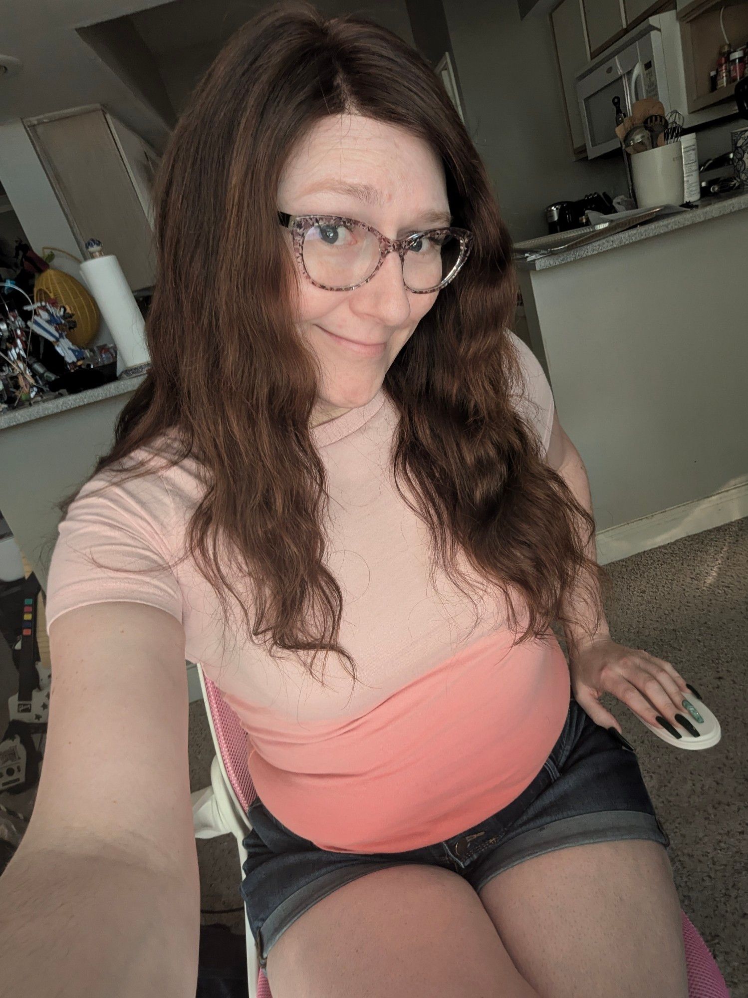 Janey sits on her pink office chair smiling wearing her trademark brown floral glasses along with a peach and pink ombre tshirt and short denim shorts.