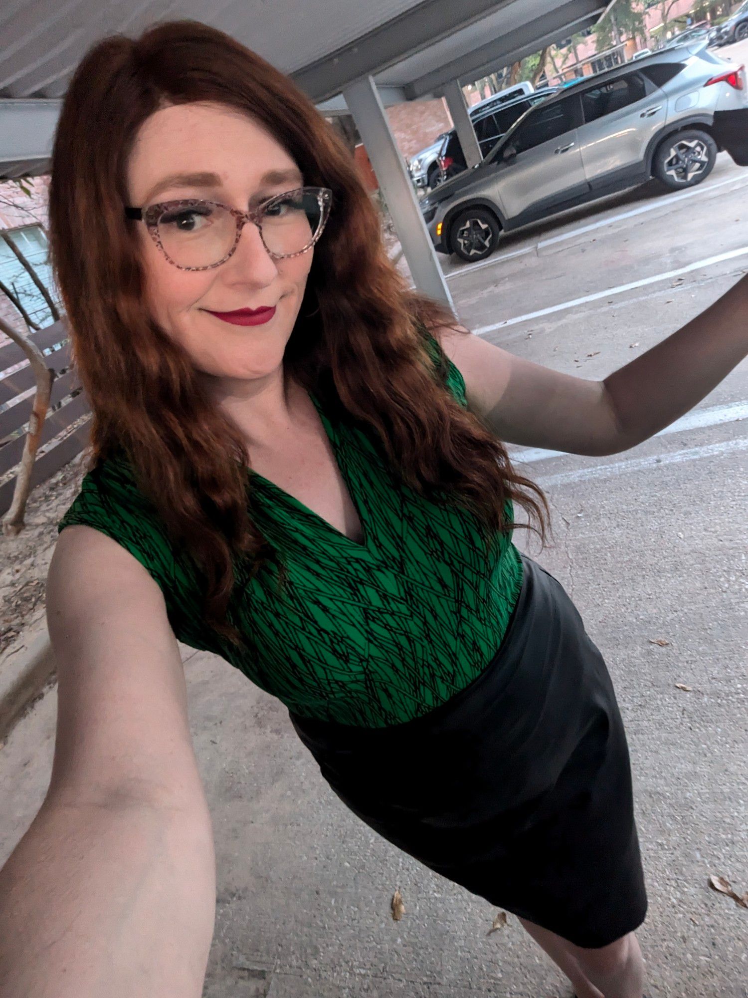 Janey stands in front of her apartment complex smiling, wearing her trademark brown floral glasses and a full face of makeup along with a green sleeveless blouse and black pleather pencil skirt.