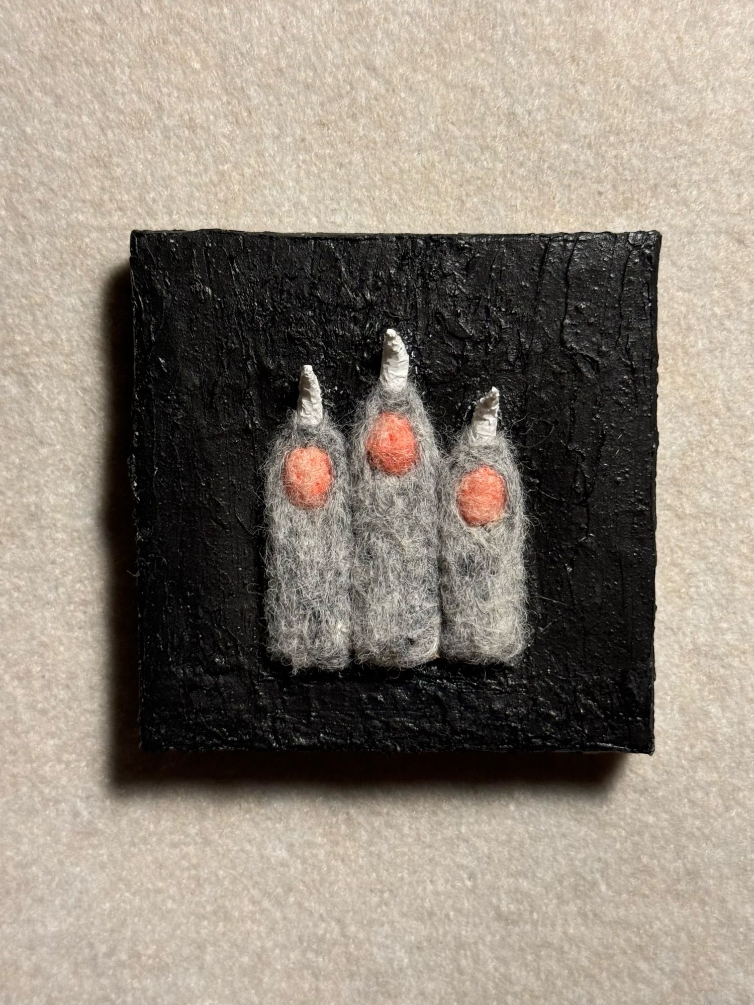 A painting on a small square stretched canvas. The background is a rough heavy body spread of graphite acrylic paint. The subject is the top half of three fingers of a creature pointing up and lined up next to each other. The fingers are gray speckled with black felted wool. The paw pads are a light pink felted wool. The claws are sculpted white acrylic