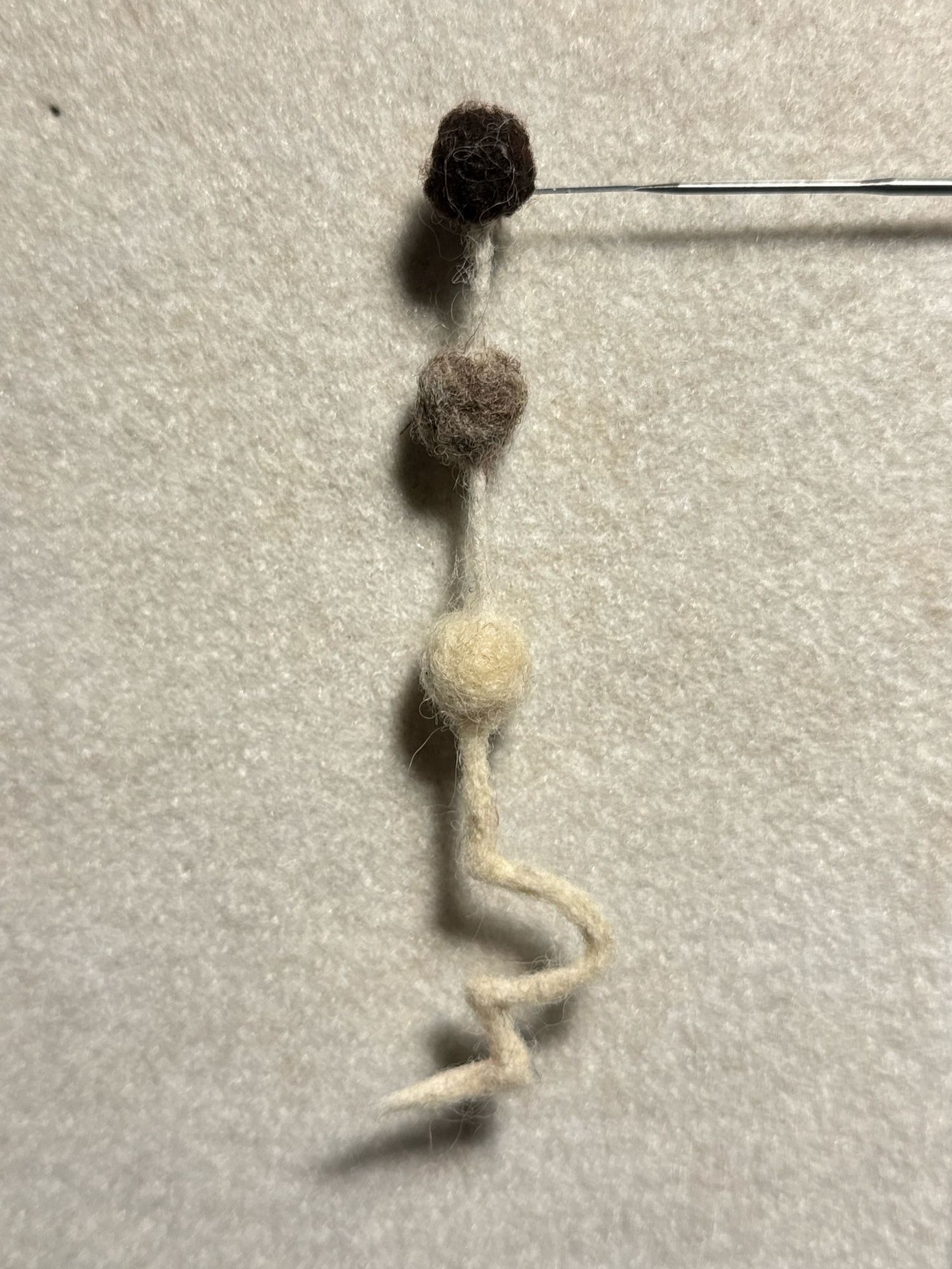 A needle felted object hanging from a felting needle in front of a beige fuzzy wall. The object is three equally sized and vertically aligned balls attached by a thin strands made of felt. Starting from the top one the color of the balls is dark brown, grayish brown, and white. The bottom of white ball has an angular and curved white line with a pointed end. A shadow of the object is cast on the wall.