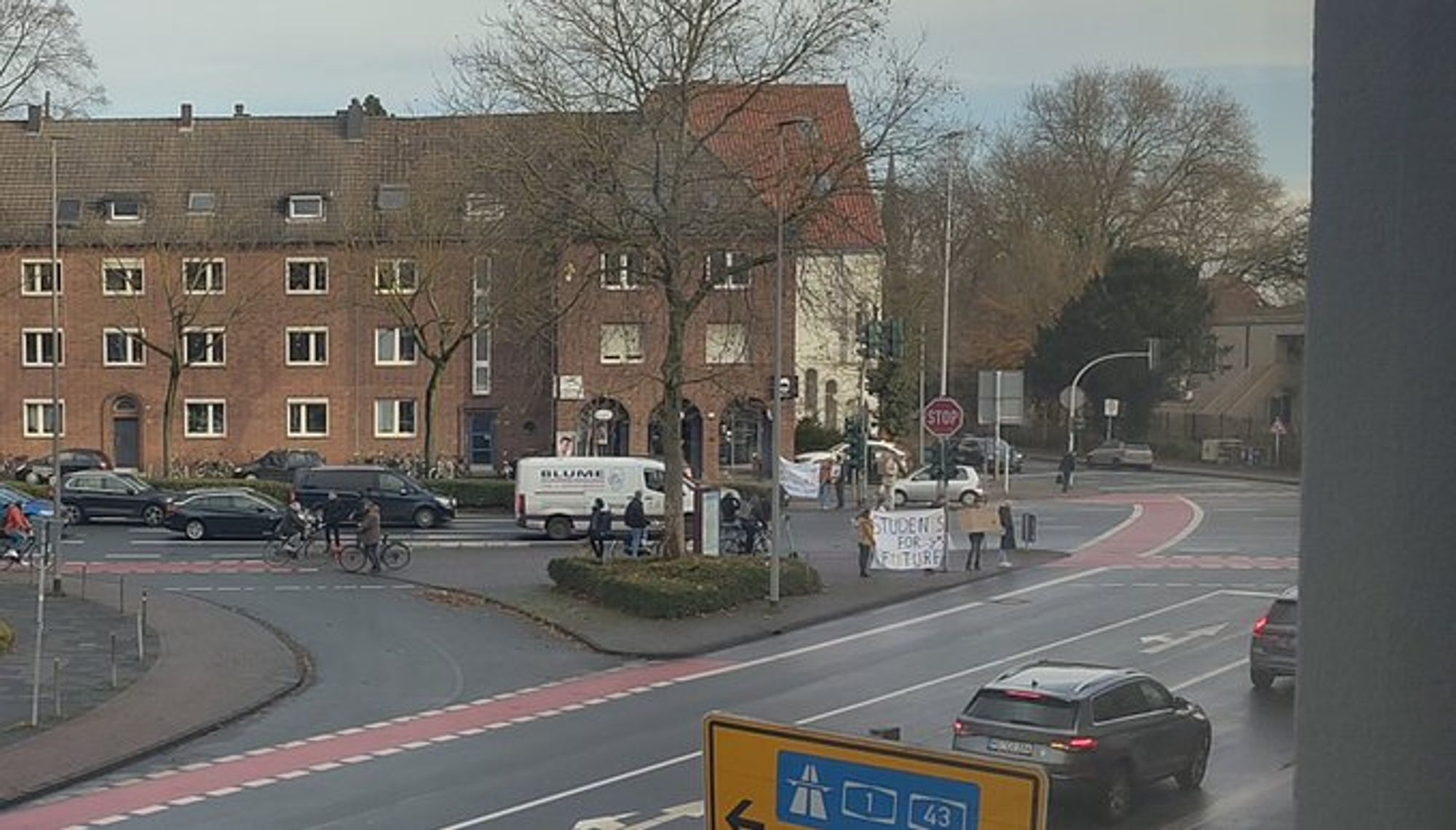 Eine Kreuzung mit einer kleinen Gruppe von Demonstrant*innen die mit Schildern und Transpis am Straßenrand stehen.