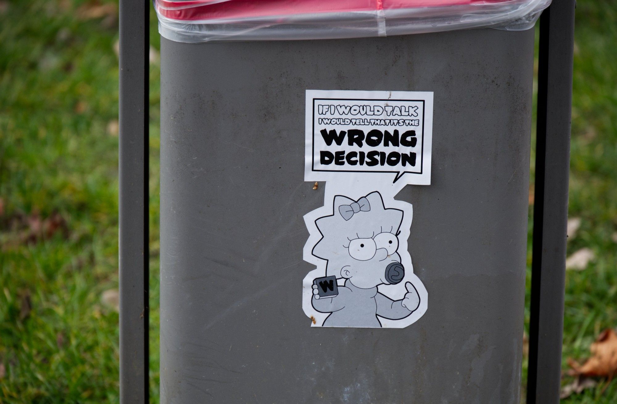 Ausschnitt zeigt einen grauen Papierkorb auf dem ein schwarzweißer Aufkleber mit der Comic-Figur Maggie Simpson geklebt wurde, die in der Spruchblase von einer "Wrong Decision" spricht.