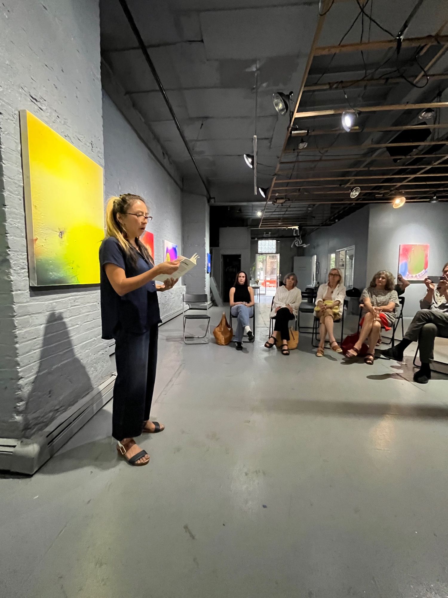 Jiwon Choi standing in front of a painting in FiveMyles reading from her book to an audience sitting in a semi circle.