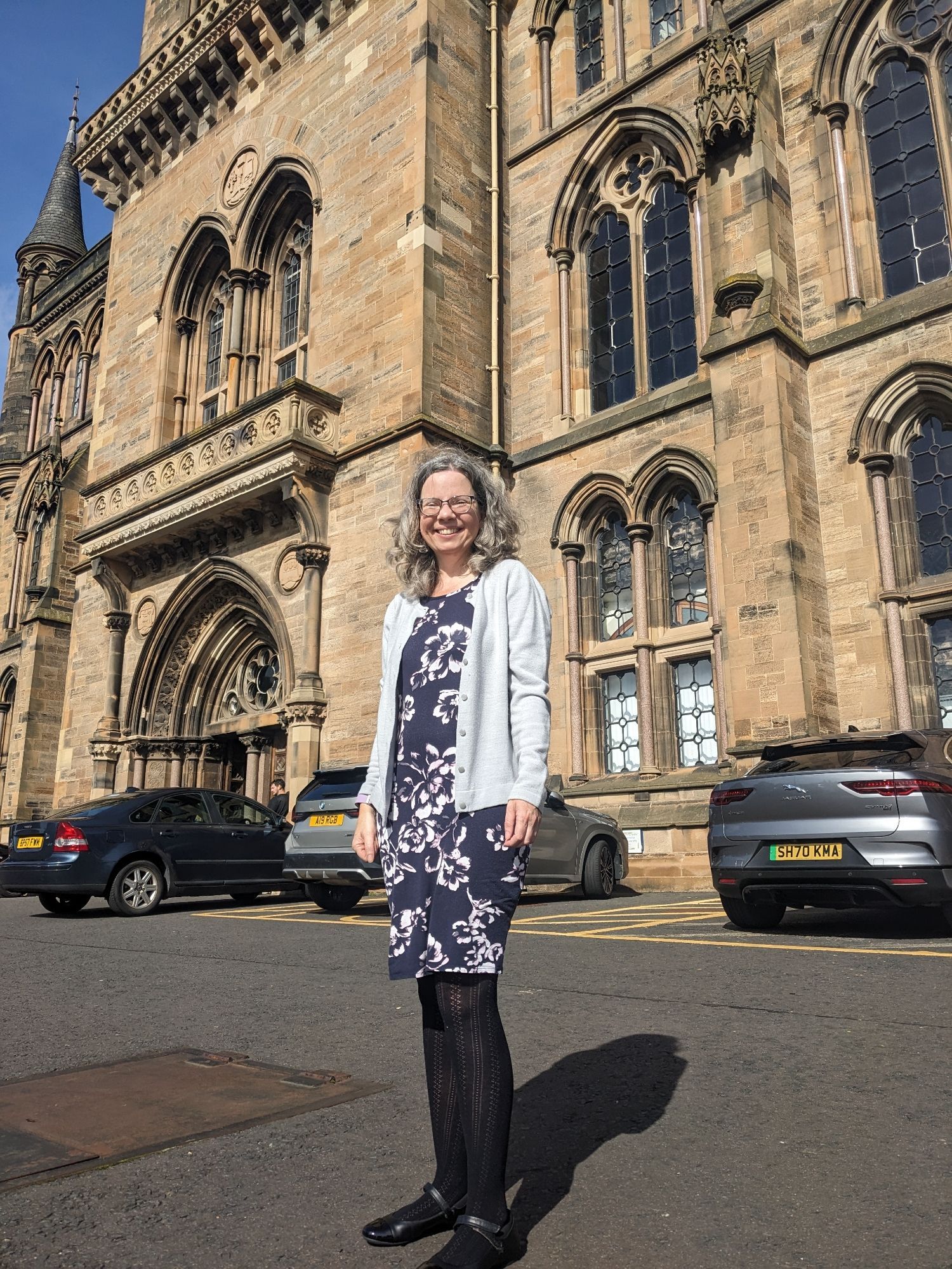 A photo of me, a white woman with curly brown hair wearing smart dress and cardigan standing in front of a Glasgow uni main building