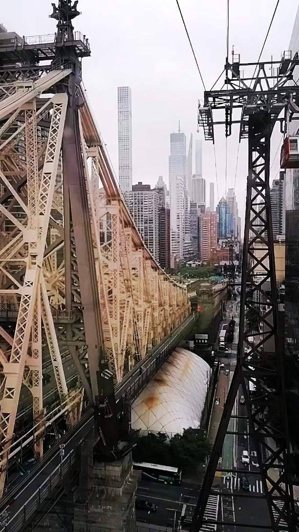 Links. Stahlgitterbrücke dazwischen unten eine Straße.. Rechts Stahlgitterpfosten einer Seilbahn. Am Horizont Hochhäuser.