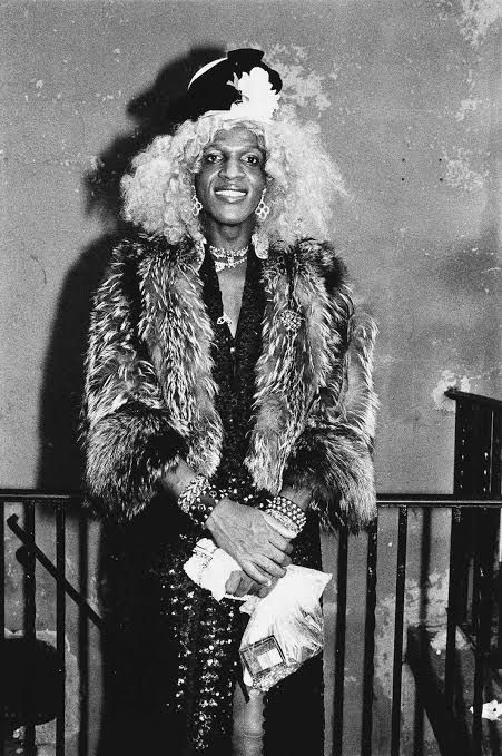 Black and white image of Marsha P Johnson. Her hair is blonde and she wears a fur coat over a sparkly black dress.