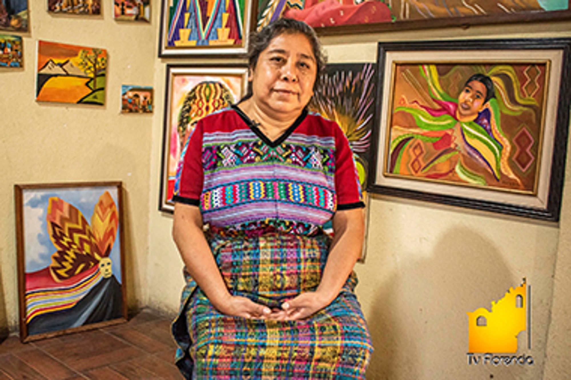 A portrait of Rosa Elena, a Maya Kaqchikel artist, in her studio. She is wearing a traditional Maya woven outfit, very colorful, and she is almost smiling for the camera. On the background are different art pieces with nature and Indigenous motifs.