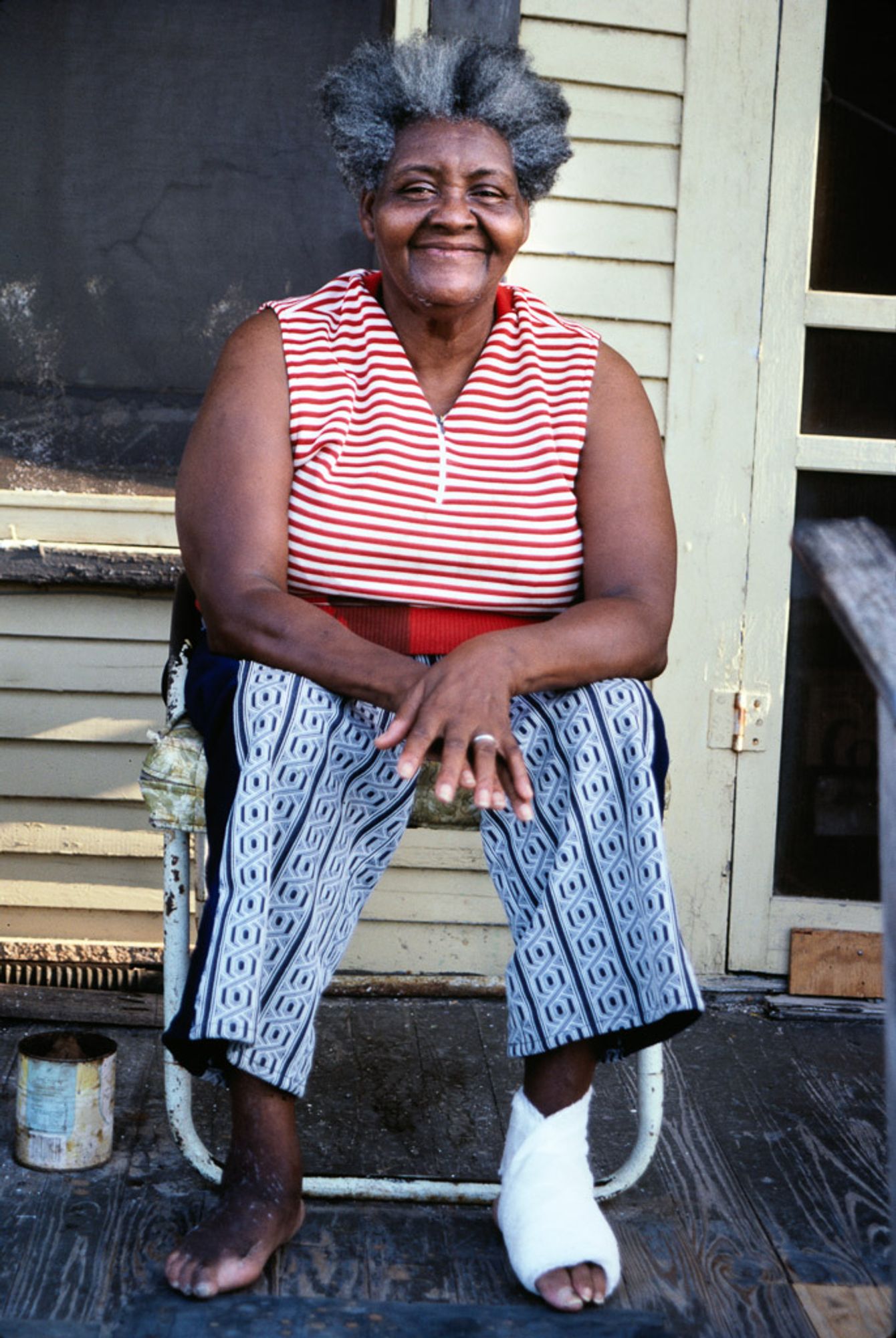 Georgia Speller (Image: William Arnett, 1987)
The first image shows an older Black woman sitting on a porch. She is smiling, wearing a red and white striped sleeveless top and black-and-white patterned pants. Her hair is gray and natural. Her foot is bandaged. The background shows a wooden house, and she sits comfortably on a chair.