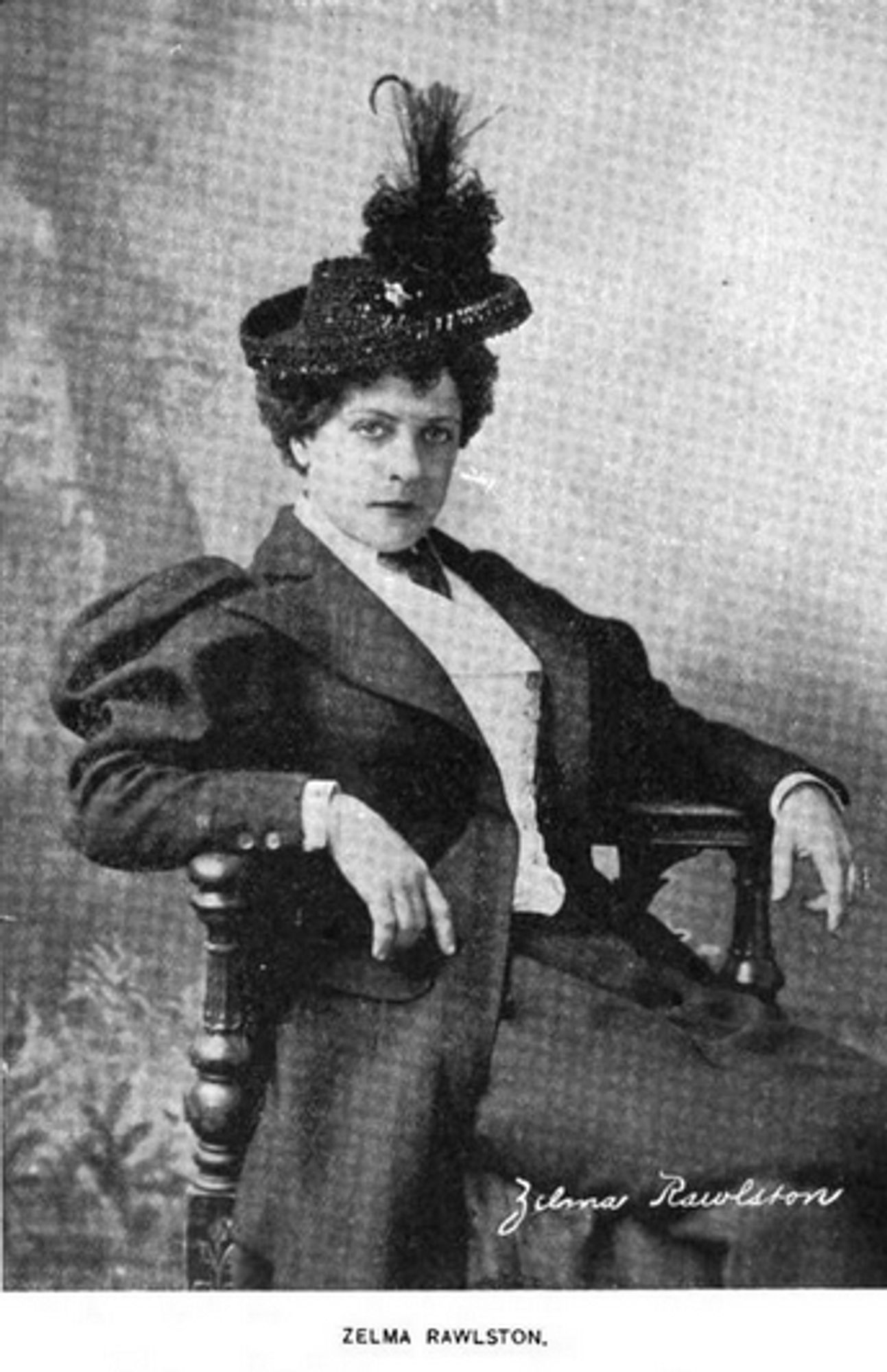 Black and white professional portrait of Zelma Rawlston seated in a high-back chair. She is dressed in a formal suit jacket with broad shoulders, a vest underneath, and a white shirt, topped off with a stylish feathered hat. She has a confident pose.