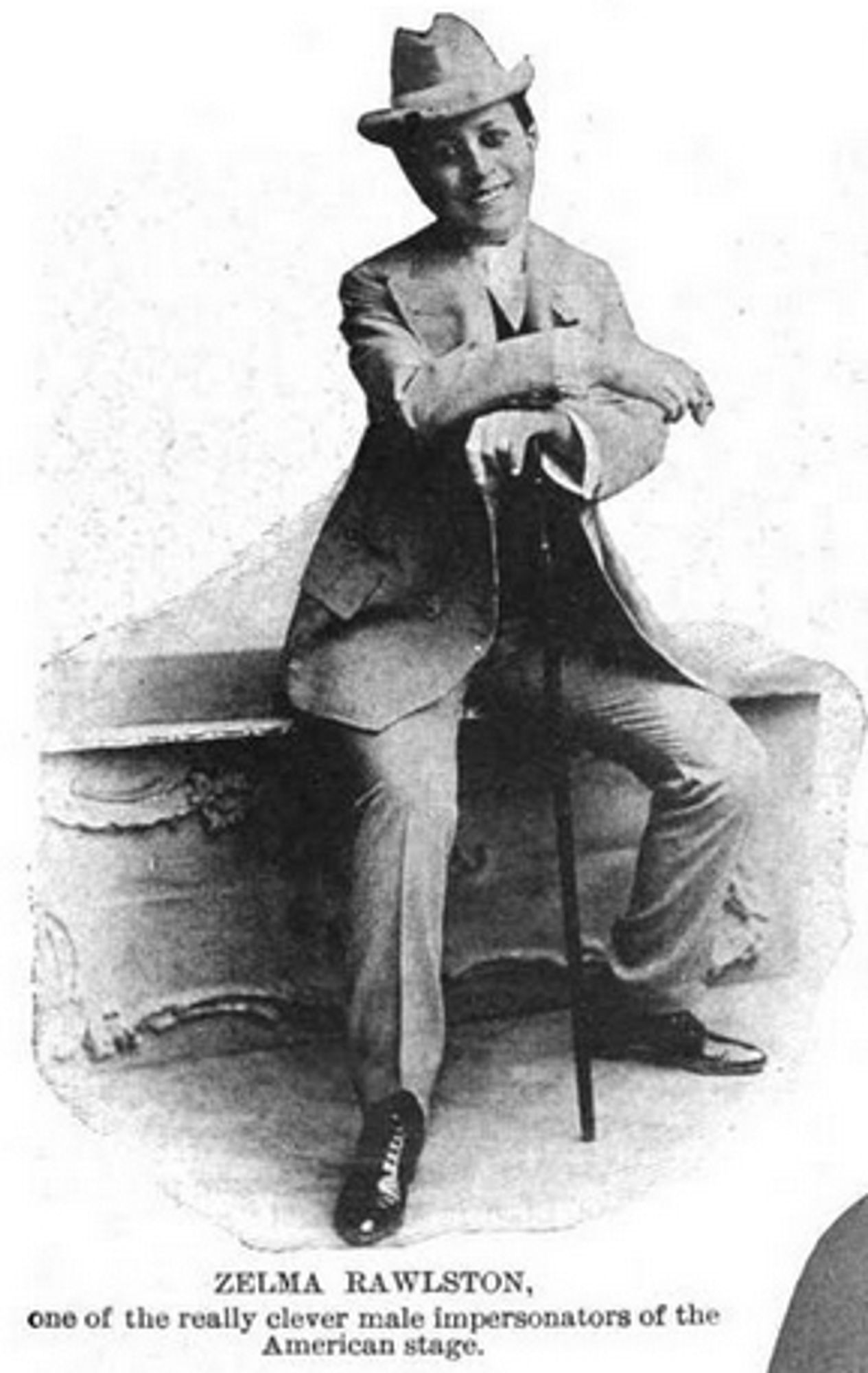 Zelma Rawlston appears in her masculine outfit, leaning casually on a walking cane. Wearing a tailored suit with a wide-brimmed hat, we see a man with ease and charm. The caption in this image highlights her as "one of the really clever male impersonators of the American stage," capturing her talent and reputation.