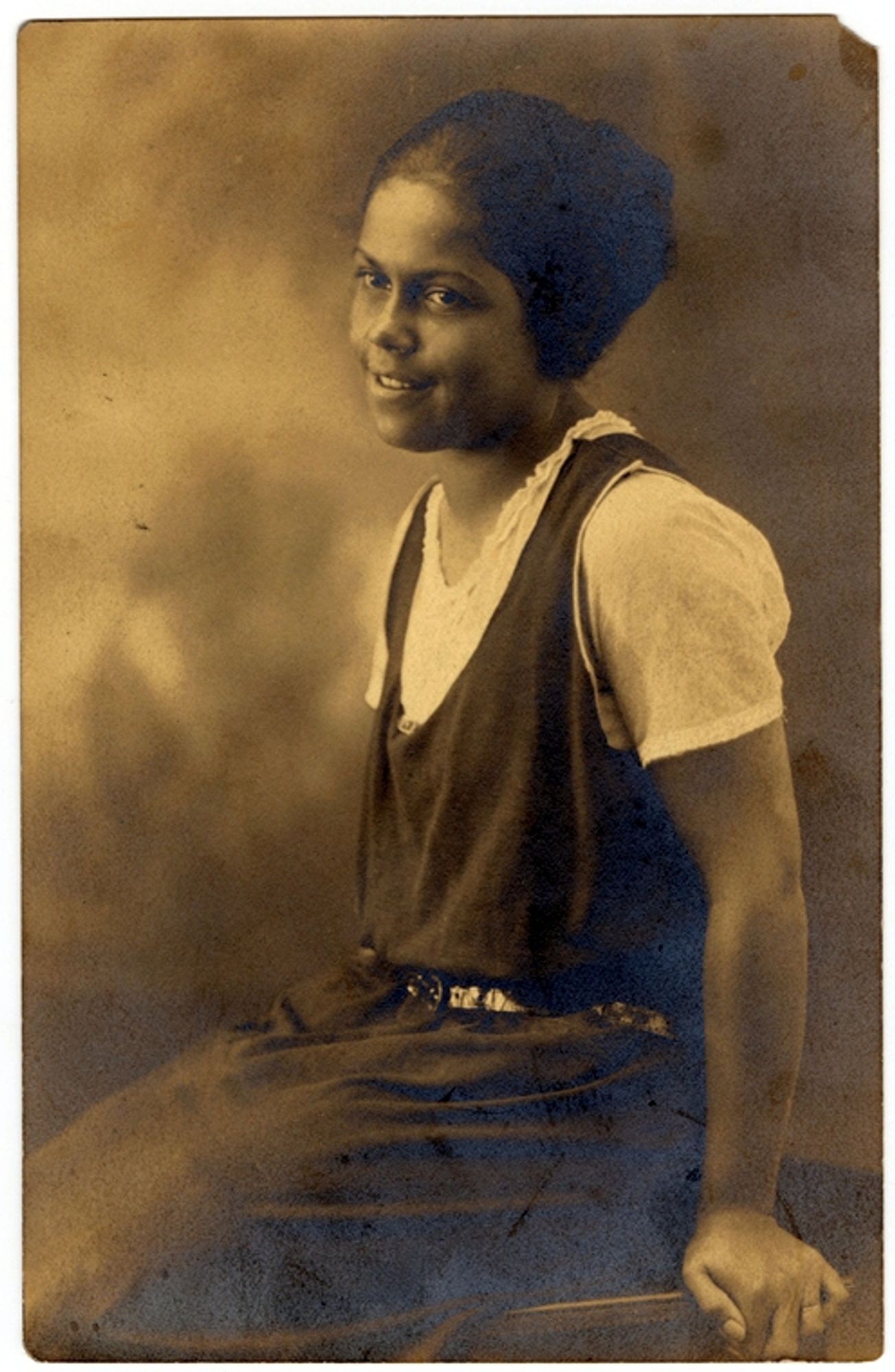 A sepia-toned professional portrait of a young Black woman, Gwendolyn B. Bennett. She is smiling gently, and she's wearing a cool outfit ahead of its time: a sleeveless, dark-colored dress over a light-colored short-sleeve top. Her hair is pulled back. The background is softly blurred, like from a photo studio.