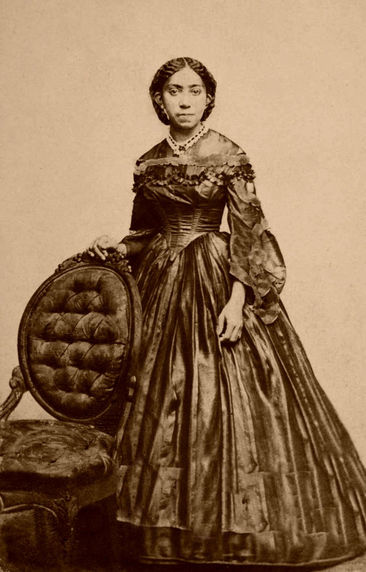 Virginia Hewlett Douglass, c. 1869. Photographed by G.H. Loomis, Boston.
Full body professional portrait. She is a young Black woman, dressed in an elegant victorian dress, looking at the camera. She is standing next to an ornate chair.