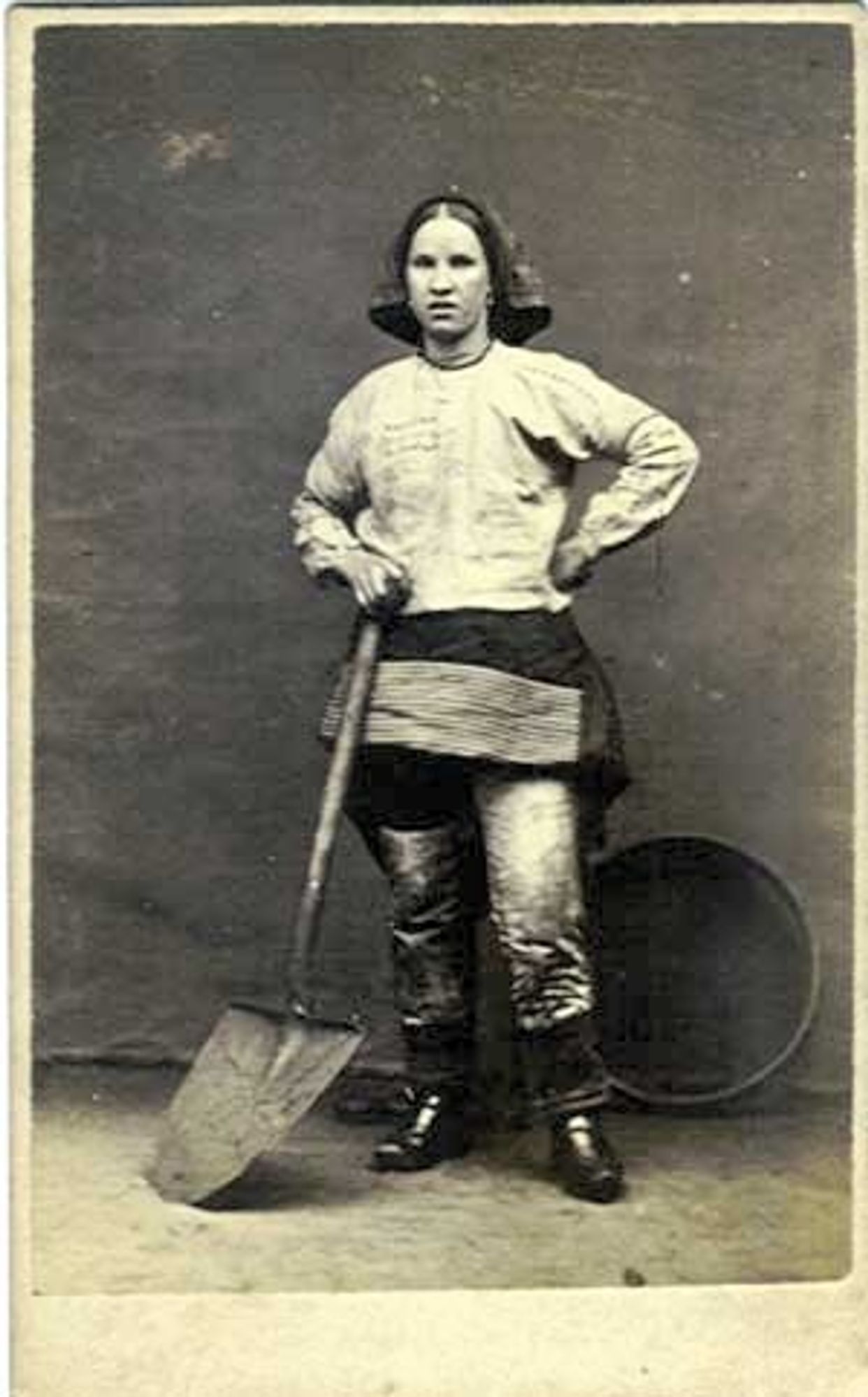 Pit brow lass from Wigan on the Lancashire Coalfield.
Black and white, full body portrait. She is a young white woman wearing miner uniform, and holding a shovel.