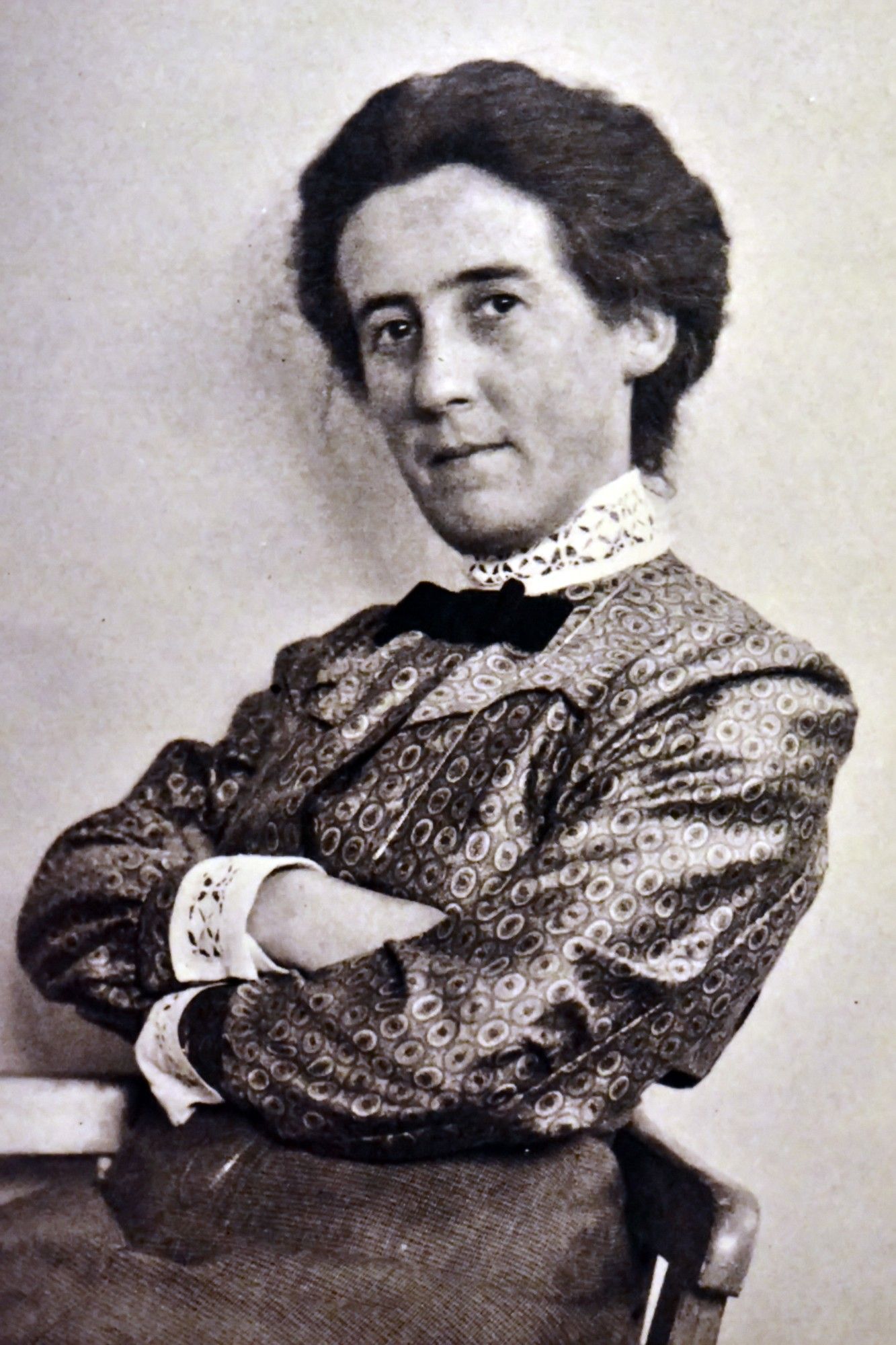 A black and white portrait of a middle aged white woman with dark hair, looking back at the camera non chalantly. She is seated, crossing her arms. She is wearing a very modern patterned blouse, adorned with white lace on the neck and sleeves.