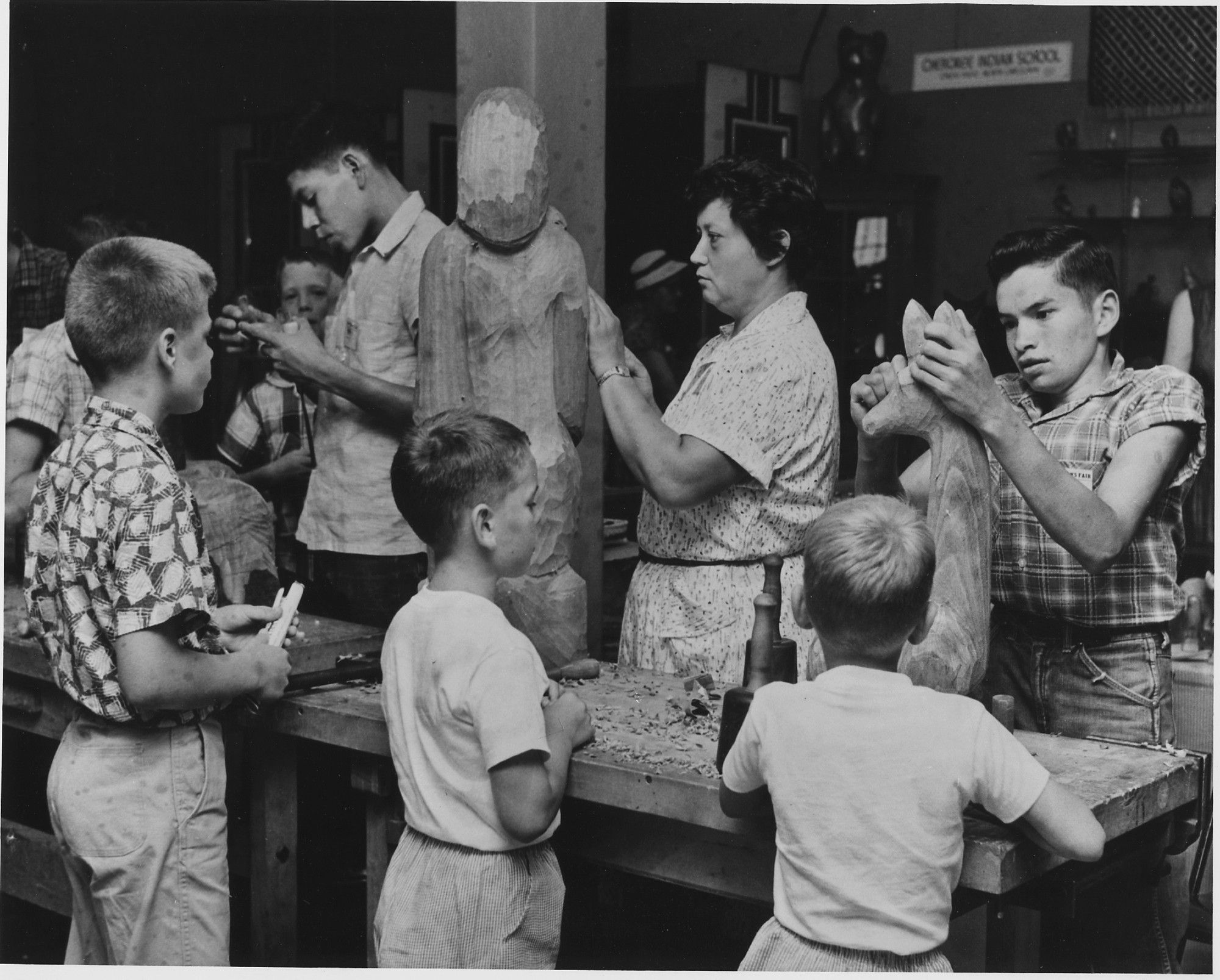 Amanda Crowe demonstrating woodcarving at the Craftsmen's Fair in Cherokee, North Carolina.
Black and white photo, showing the artist, a middle aged woman, at the middle, with a big wooden piece being carved. She is surrounded by children of different ages, each focused either on their own carvings or her demonstration.