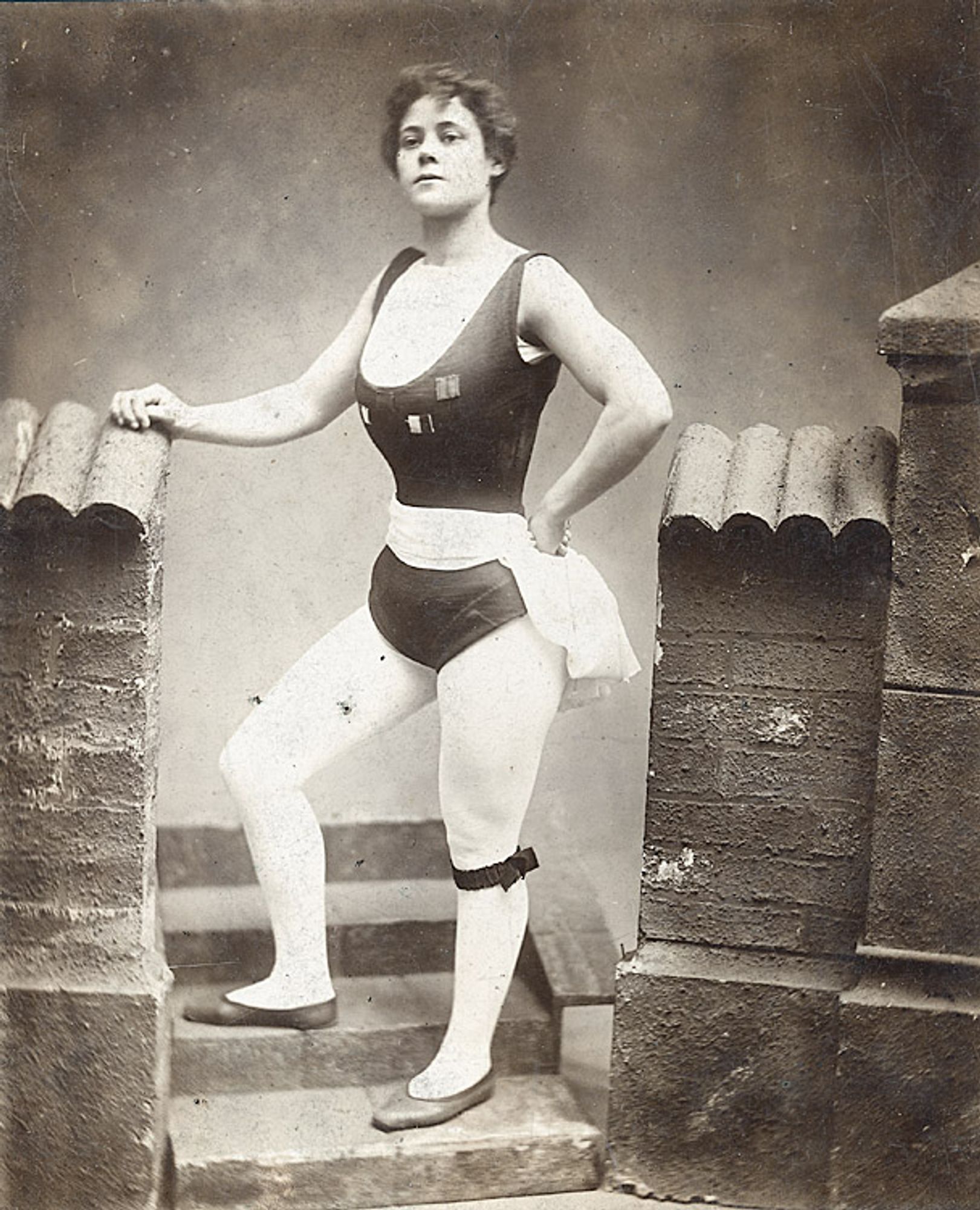 Black and white full body portrait of Vulcana, a renowned strongwoman. She is a young, powerful white woman who stands confidently in a classical pose. She is dressed in a sleeveless athletic outfit that accentuates her muscular physique.