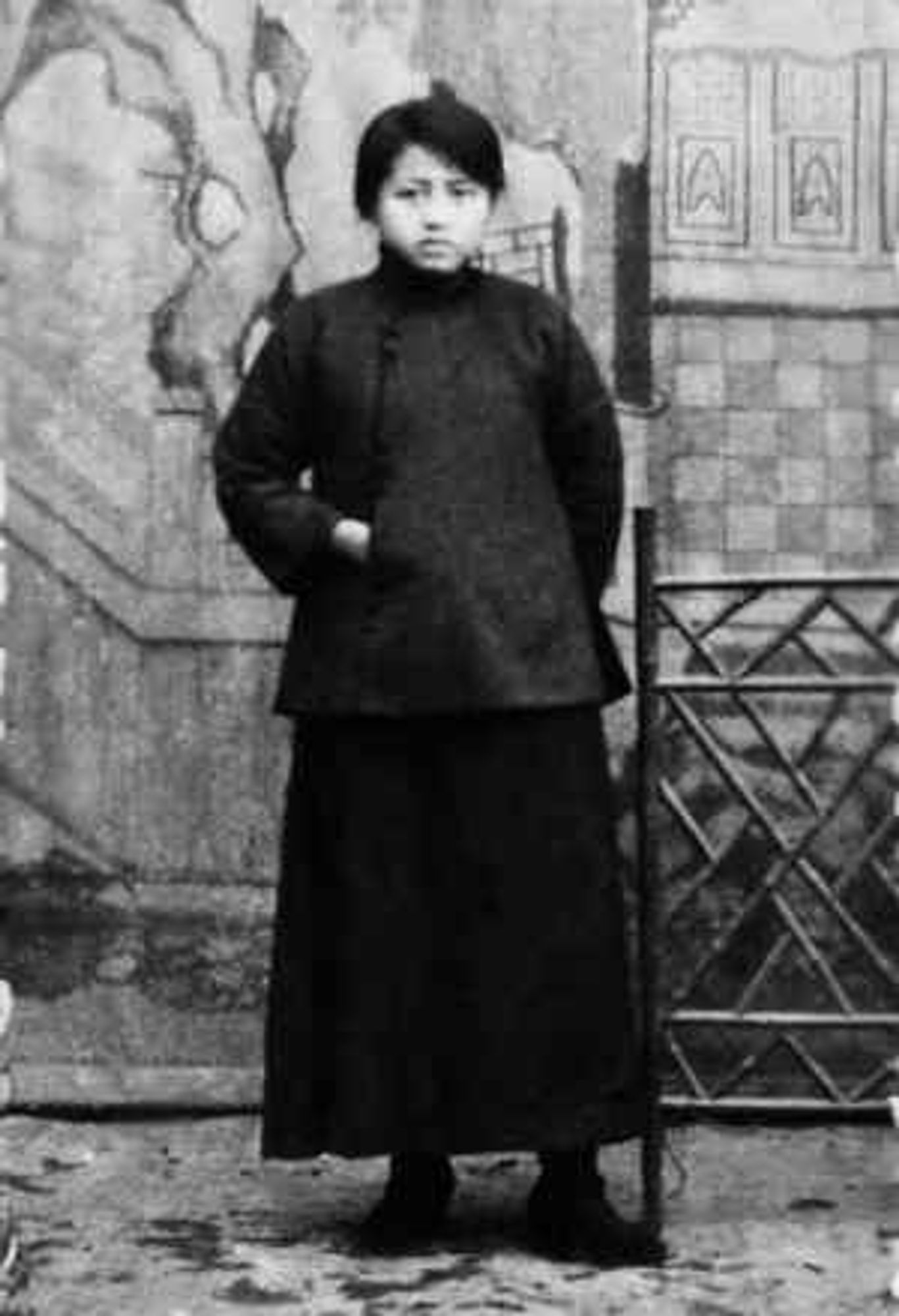 A full body black and white portrait of Wang Huiwu, a young Chinese woman standing upright in a simple black traditional Chinese garment with a serious expression. Her hands are tucked in the pockets of her jacket. The backdrop behind her seems to be a painted scene, like a photo studio.