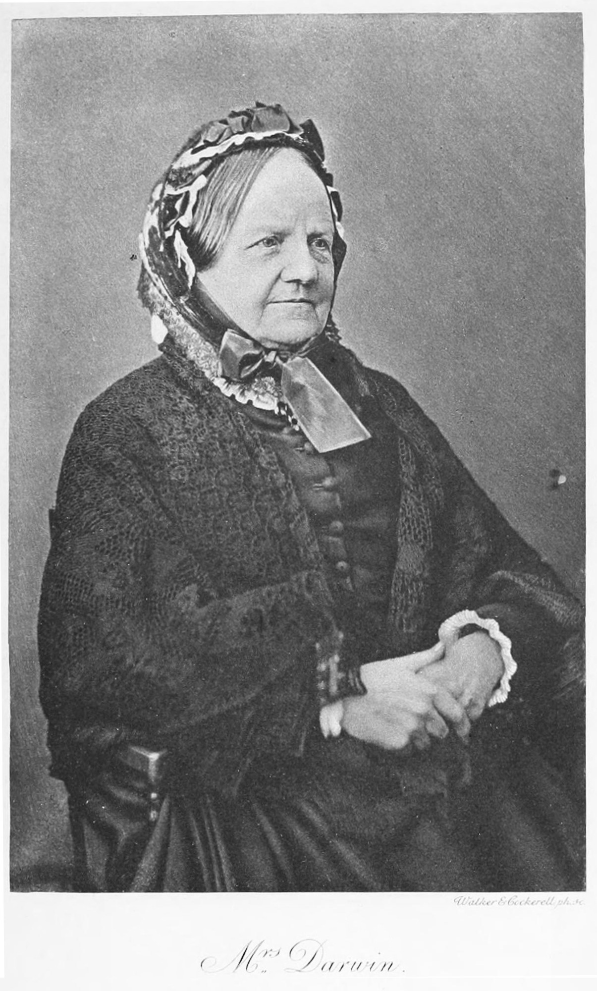 Another black and white portrait of Emma Darwin posing seated. She is older in this picture, posing with the same calm expression. She is wearing a head covering adorned with ribbons, and a modest dress and lace cape.