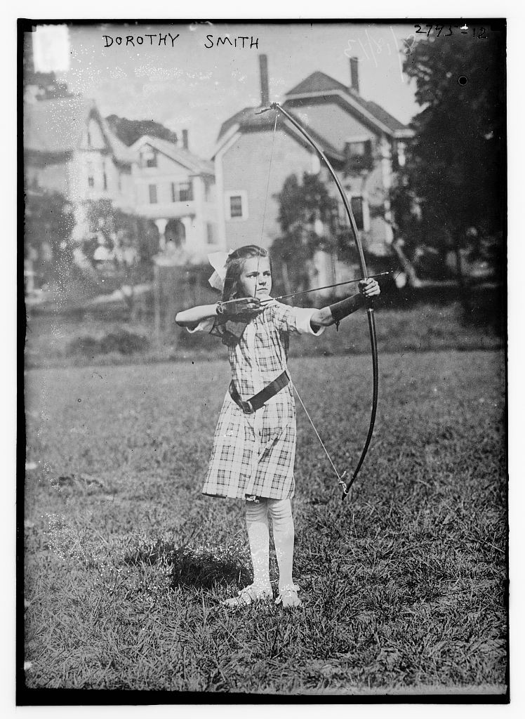 A black and white photo of Dorothy Smith in 1913 ready to shoot an arrow. She is a 10 year old girl, holding the bow and arrow like a badass. She is at a field or park on a sunny day.