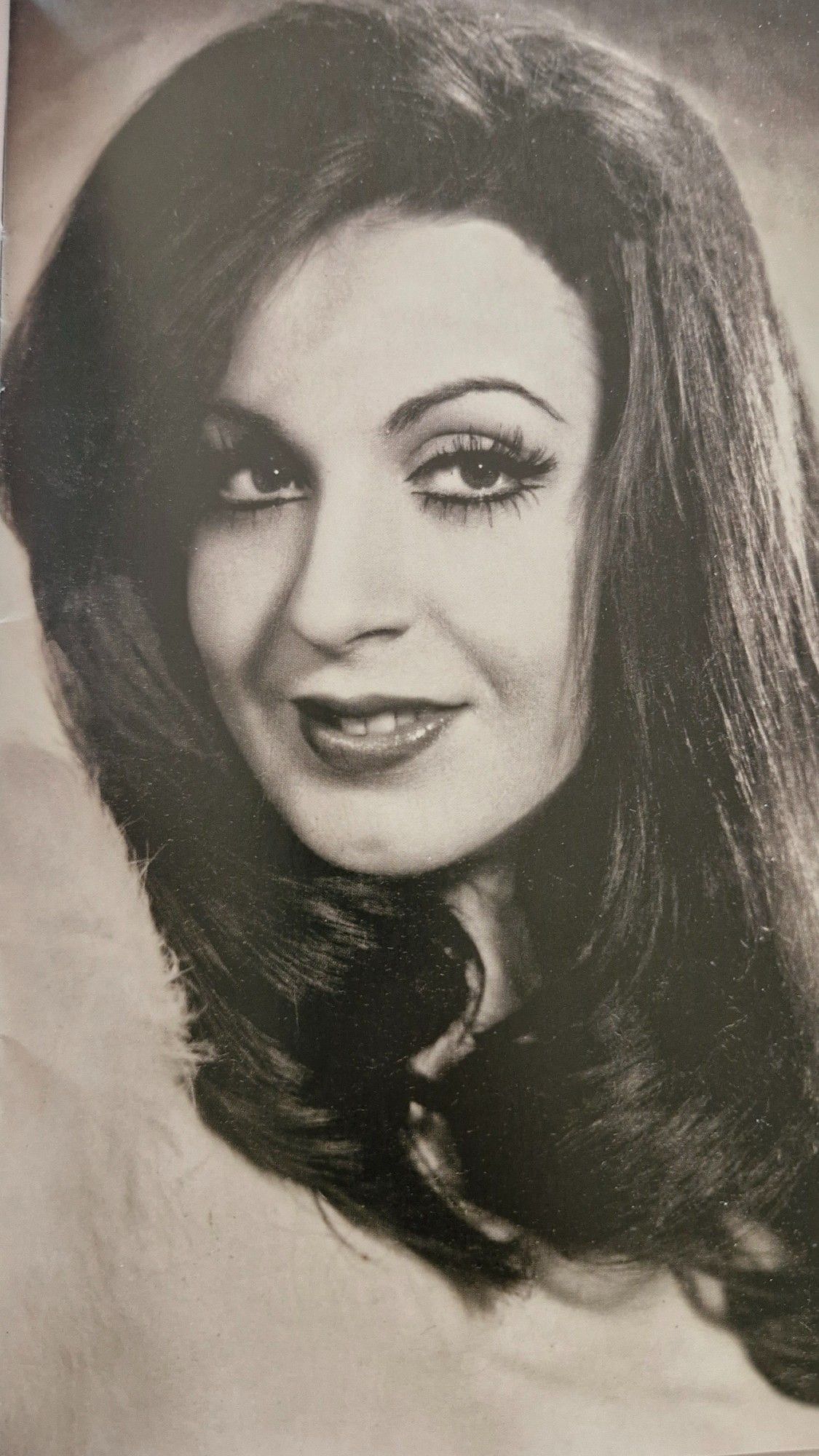 Black and white portrait of Sema Yıldız from the early 1970s. She is depicted in a close-up shot with her hair styled in soft waves. Her makeup is accentuated, with bold eyeliner and long lashes, highlighting her expressive eyes. She is smiling subtly, exuding a sense of elegance and charm.