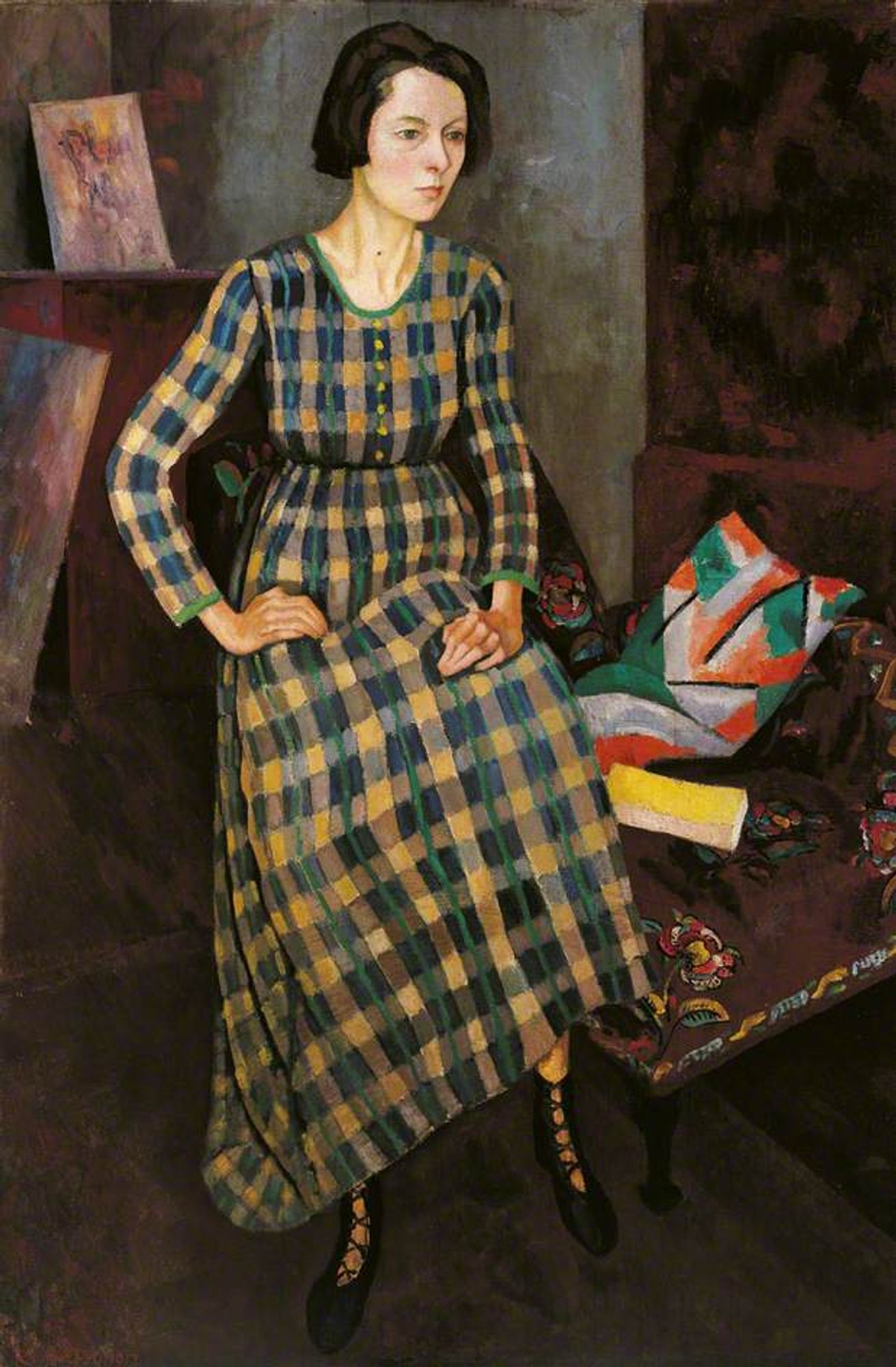 A full-body portrait of Nina Hamnett seated on a chair, wearing a long, checkered blue and yellow dress with lace-up shoes. Her posture appears thoughtful, with her hands resting on her lap, and a serious expression on her face. In the background, there are paintings and a funky cushion.