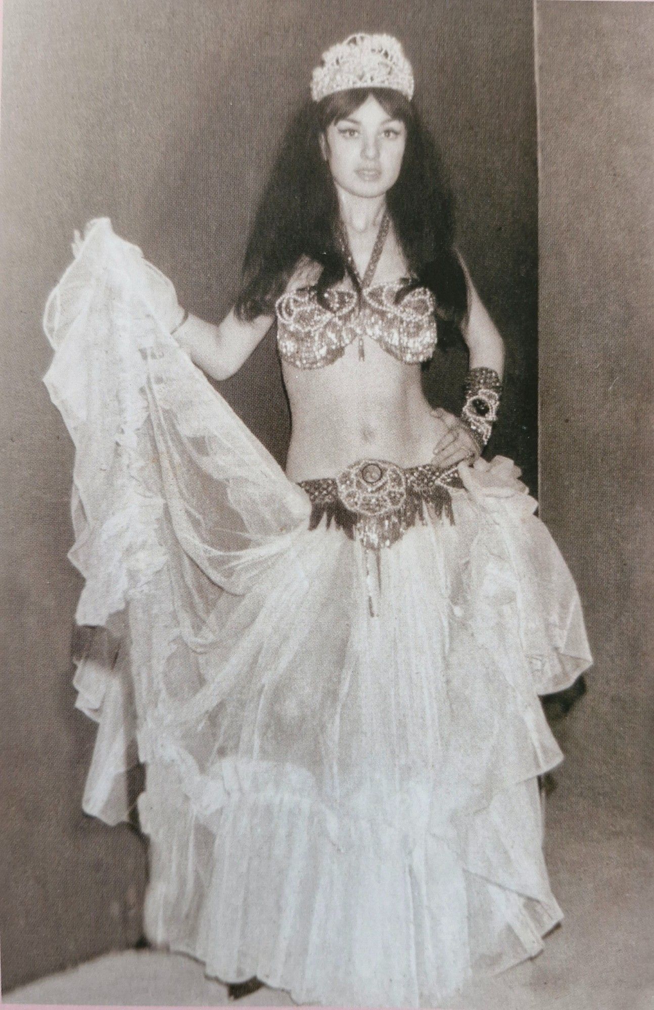 A black and white, full body portait of Sema Yıldız, a famous Turkish belly dancer, in 1967. She is dressed in a traditional belly dancing costume, adorned with intricate beadwork and accessories. The costume features a jeweled bra, a belt, and a flowing skirt. She is posing with one arm raised, holding a part of her skirt.