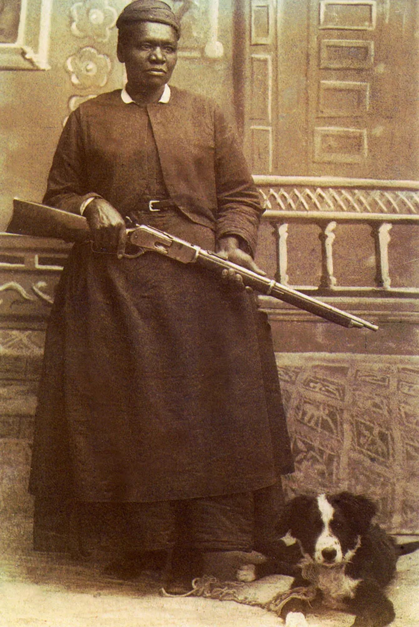 Sepia coloured portrait showing a Black woman standing confidently, dressed in a practical long dress, holding a rifle. Her posture and expression convey strength and determination. A dog (medium, black and white, kind of like a Sheppard dog) sits next to her. The background features painted ornate architectural details, like a stage for a photo studio.