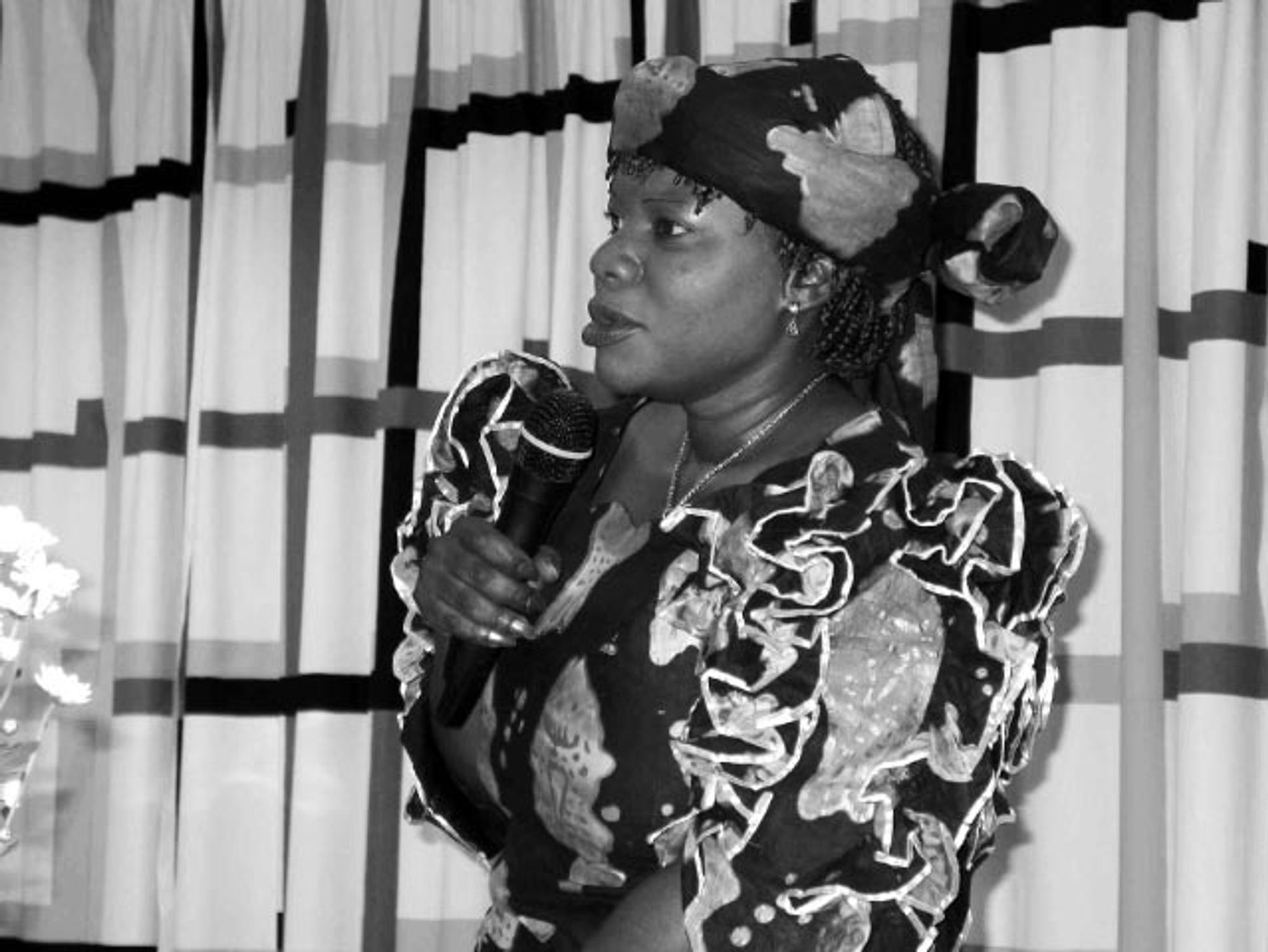 A black-and-white photograph of Margared Dongo, a middle aged Black woman, standing and speaking into a microphone in what looks like an official political event. She is dressed in a nice traditional African outfit decorated with fish pattern, and a headscarf. Her dress is also adorned with ruffled sleeves. There is a backdrop of curtains behind her.