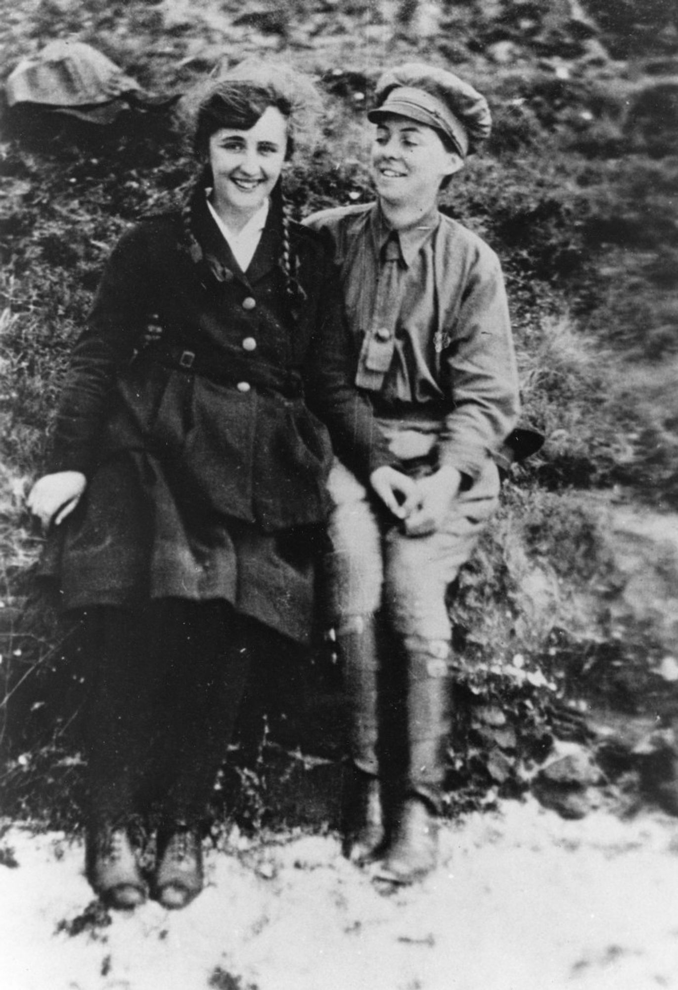 This is a black-and-white photograph of two women sitting outdoors, both smiling. The woman on the left is wearing a dark, belted coat and has braided hair, and she's looking at the camera while the woman on the right, Anderson, is wearing a masculine outfit that includes a hat, a tie, and long boots. They are laughing, seated close to each other on grass or rough terrain, holding hands. The backdrop is a natural setting.