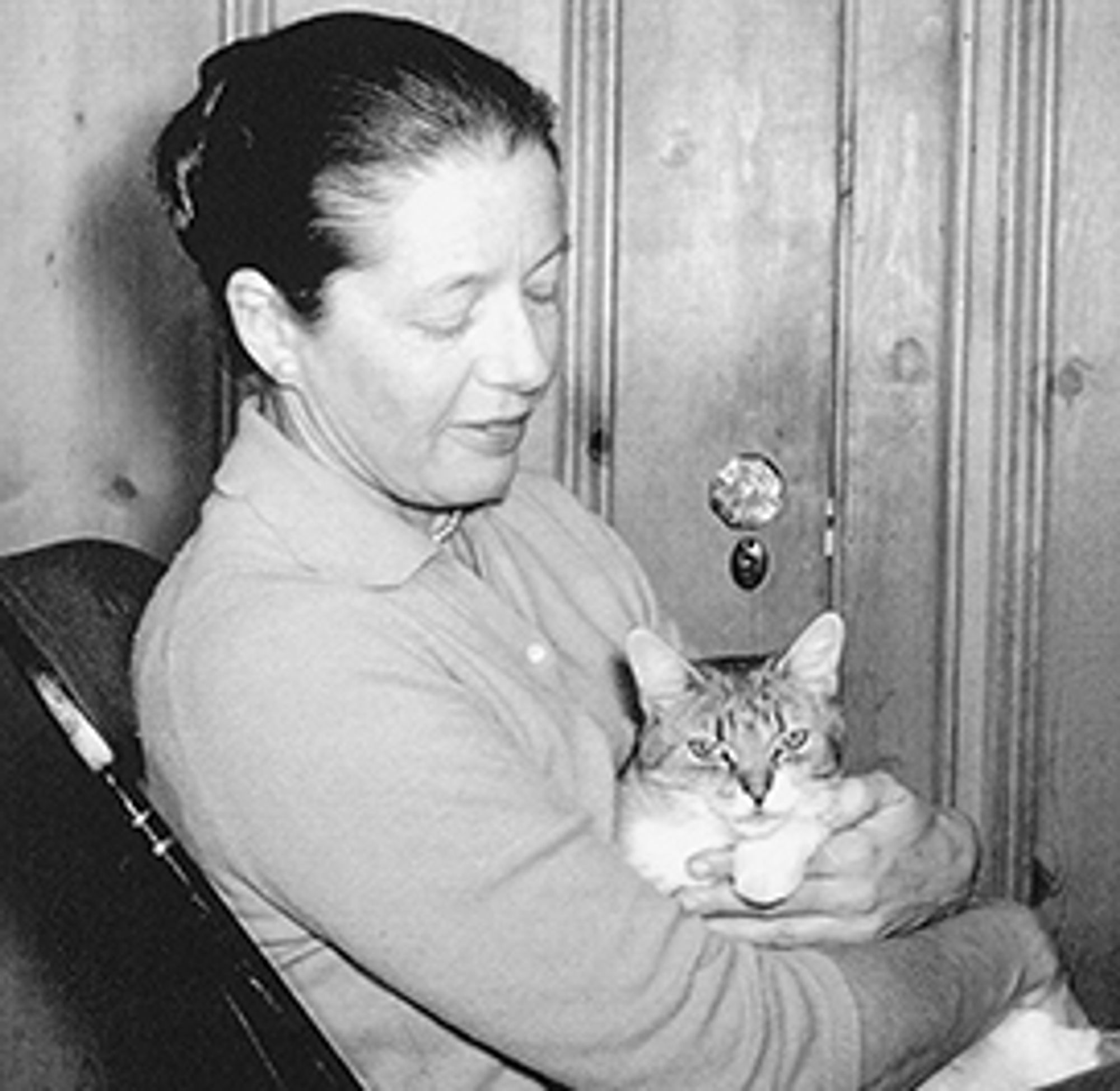 A black and white photo of Jean Holzworth, a middleaged white woman, siting and holding a cat in her arms. The cat is stripy, and looking at the camera, maybe with a bit of distrust to the photographer. The doctor is looking at the cat with an expression of care.