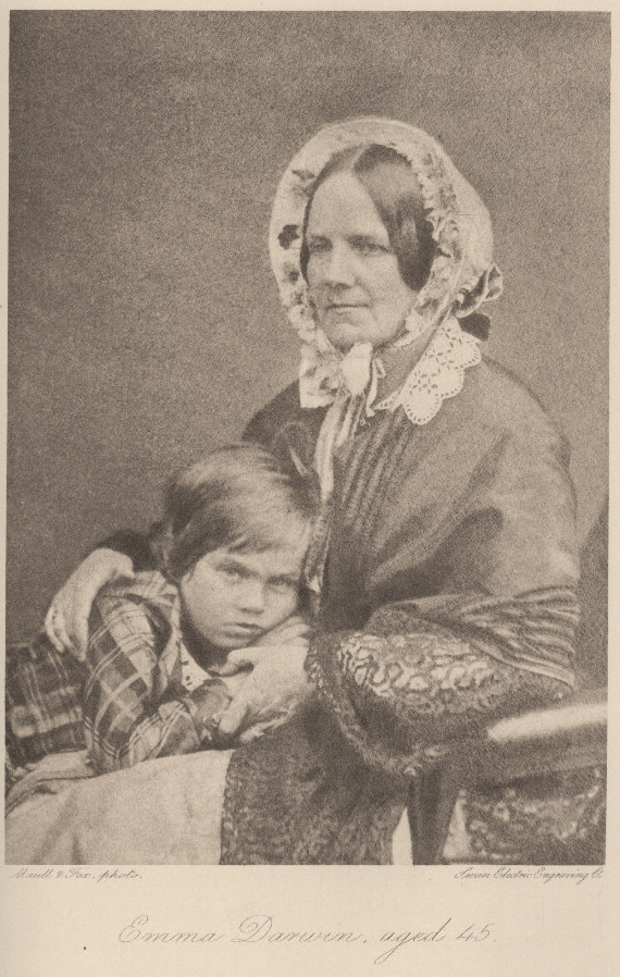 A black and white photo of Emma Darwing with a young boy on her lap that she is hugging. The boy looks at the photographer with distrust, while Emma poses calm. The photo is printed, ant it reads at the bottom with cursive lettering "Emma Darwin, aged 45".