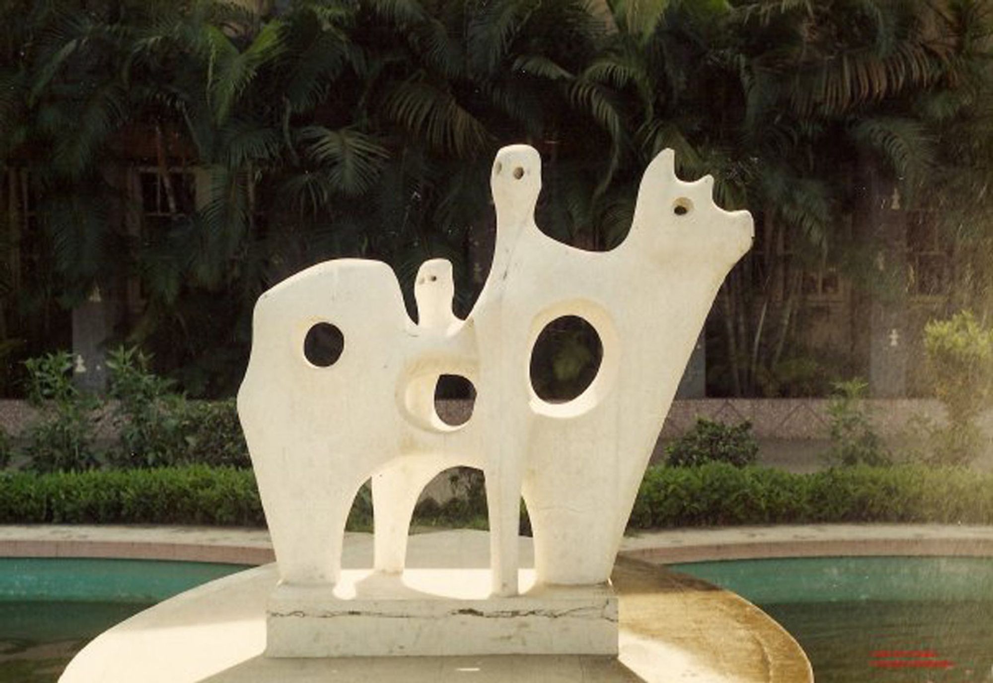 A minimalist marble sculpture titled "Cow with Two Figures" created by Novera Ahmed in 1958. The abstract shapes a cow with two humanoid shapes interacting with her. The sculpture has smooth, curved lines and round cut-out holes, blending them together. It is set outdoors with lush greenery in the background.