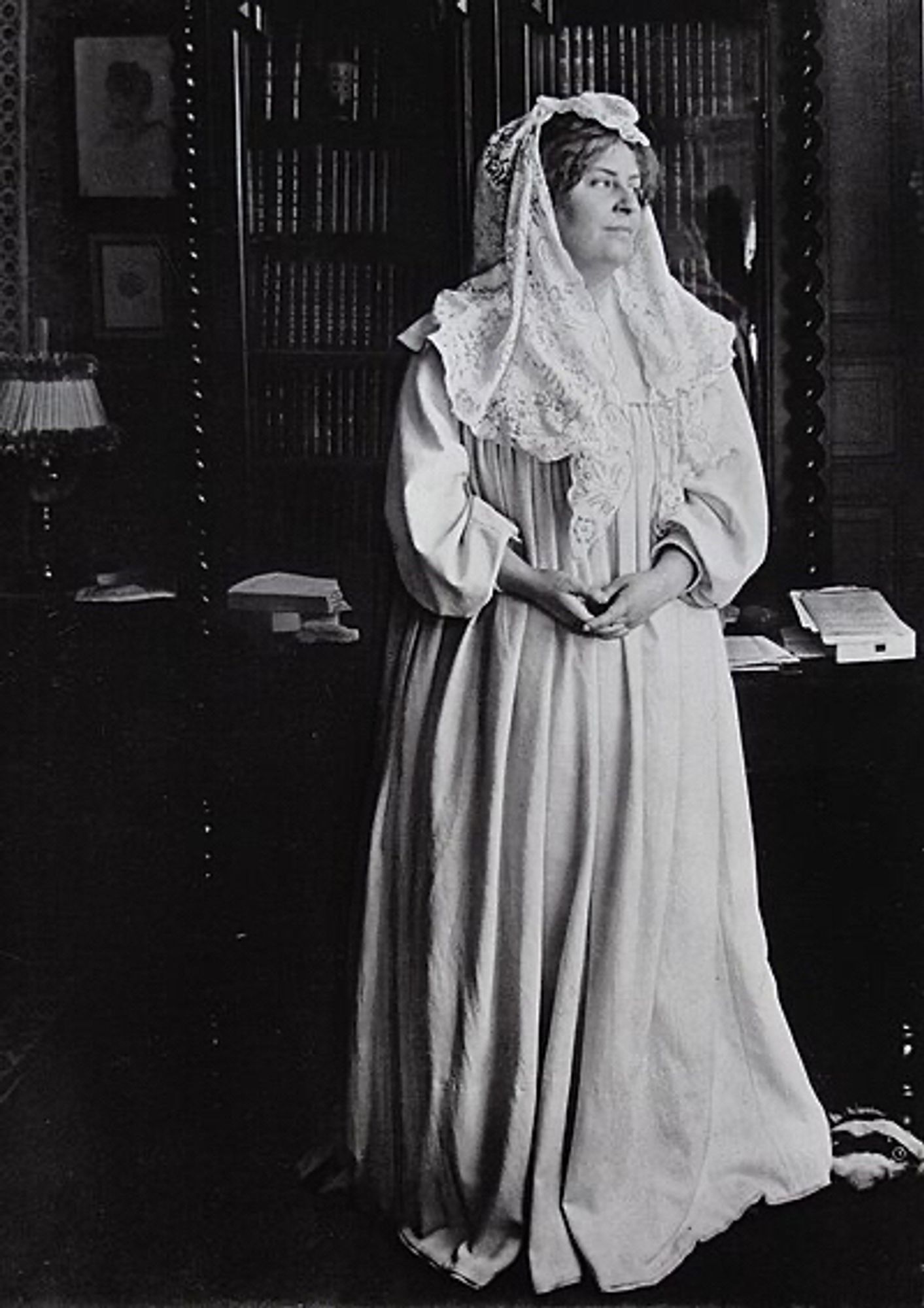 Full body black and white portrait of a white woman standing at a library. She is wearing a white dress and veil. She is looking to the side.