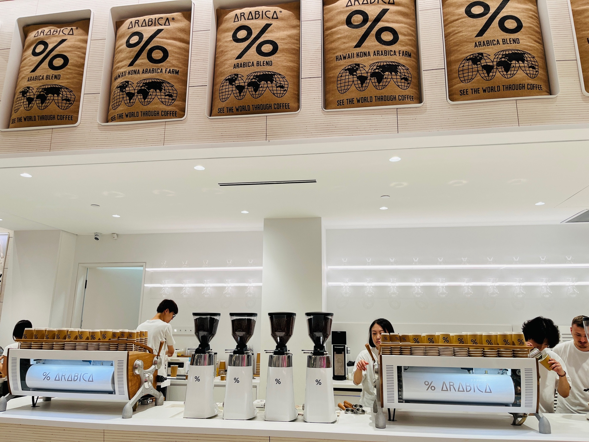 Photo of a Minimalist designed coffee shop. With large coffee bags on top in a row for design. And Baristas brewing coffee behind coffee machines. 