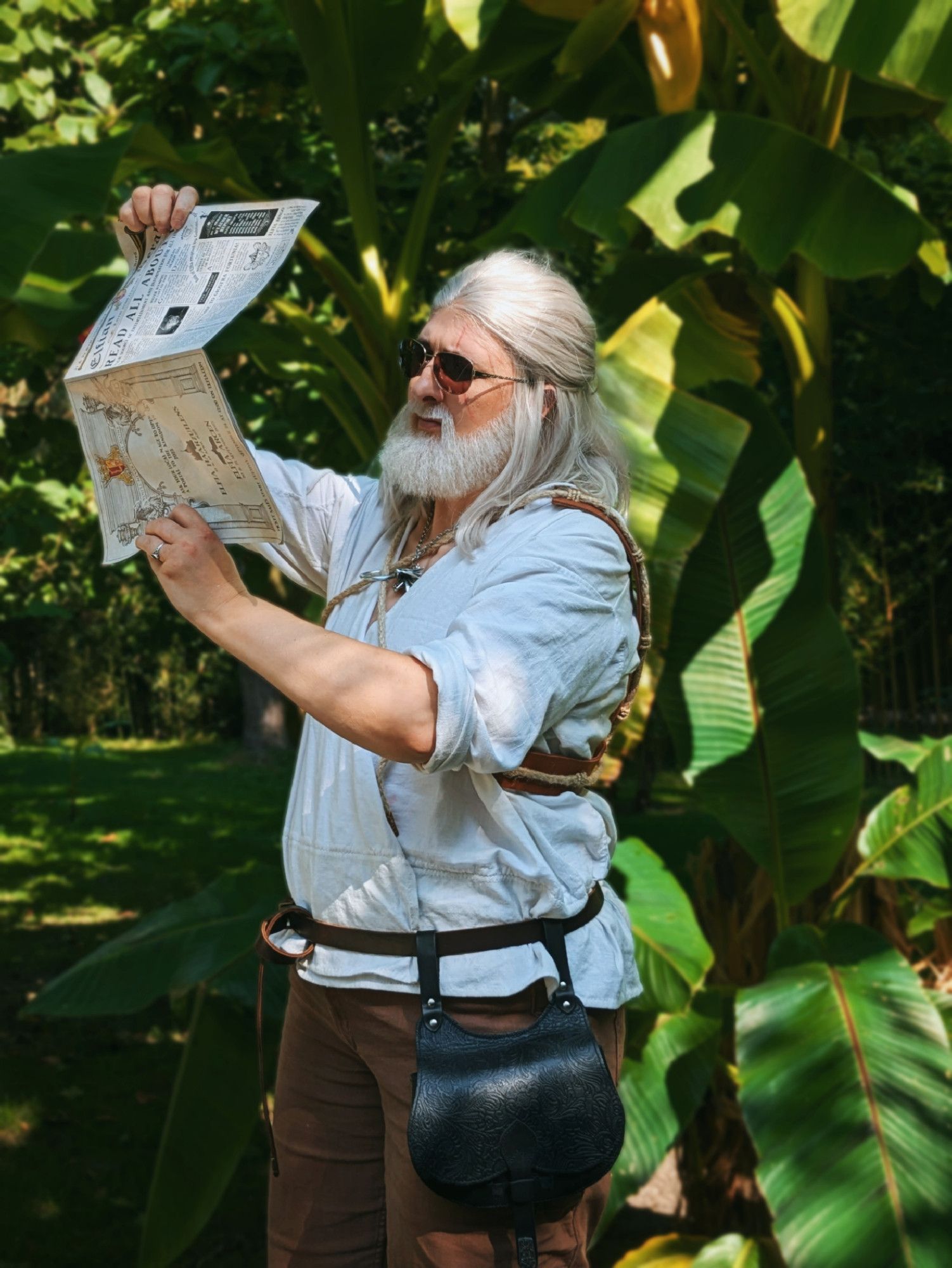 Geralt cosplayer from the Witcher WIld Hunt standing next to palm trees holding up a leaflet in entirely the wrong way. He's wearing sunglasses and seems ready for vacation but is obviously quite lost.